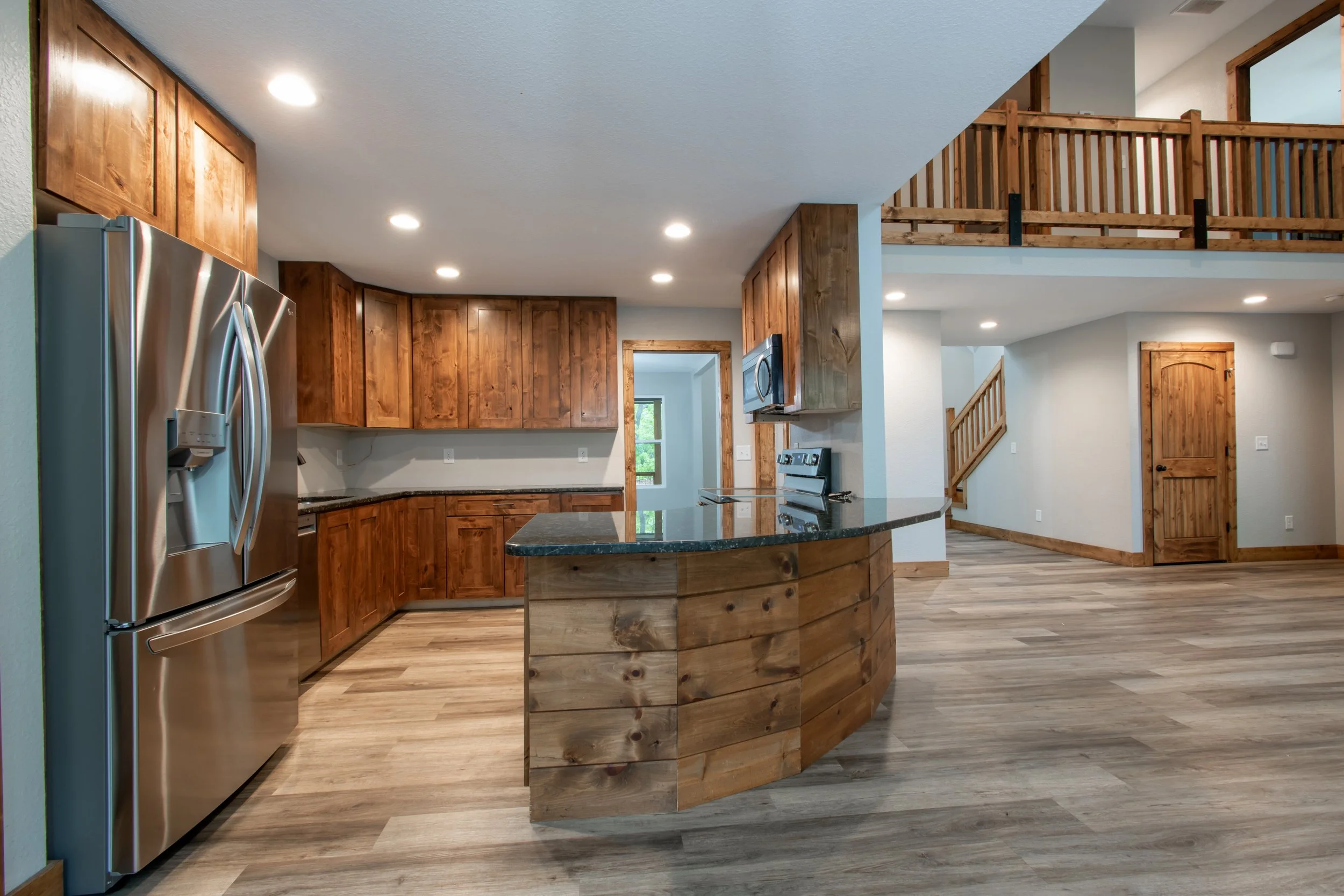 Open-plan kitchen and living area with wooden cabinets, a stainless steel refrigerator, a black granite counter island, and wooden features including a staircase and railing, with hardwood floors and recessed lighting.