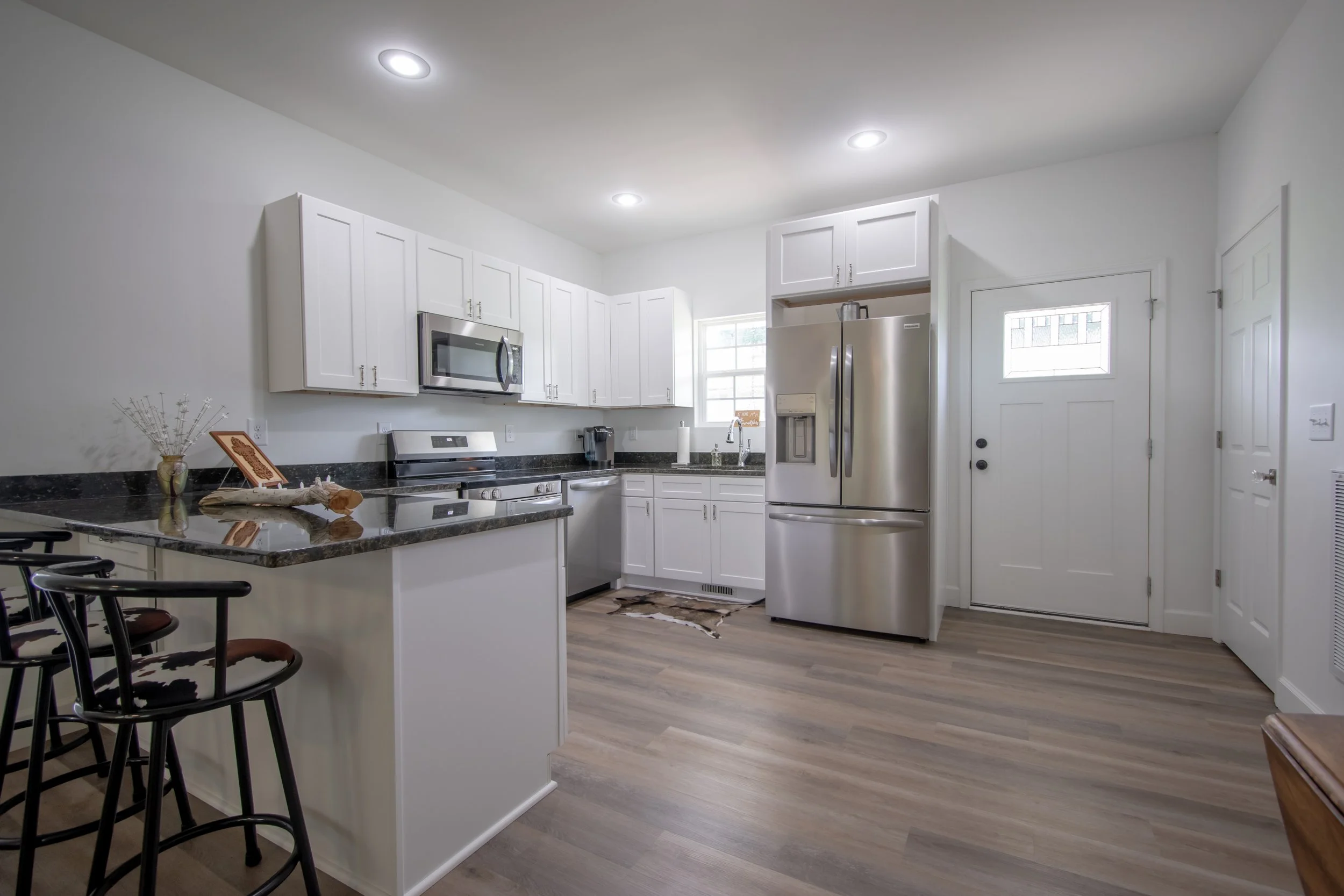 Modern kitchen with white cabinets, stainless steel refrigerator, microwave, and stove, with wood laminate flooring, a small window, and a white door.