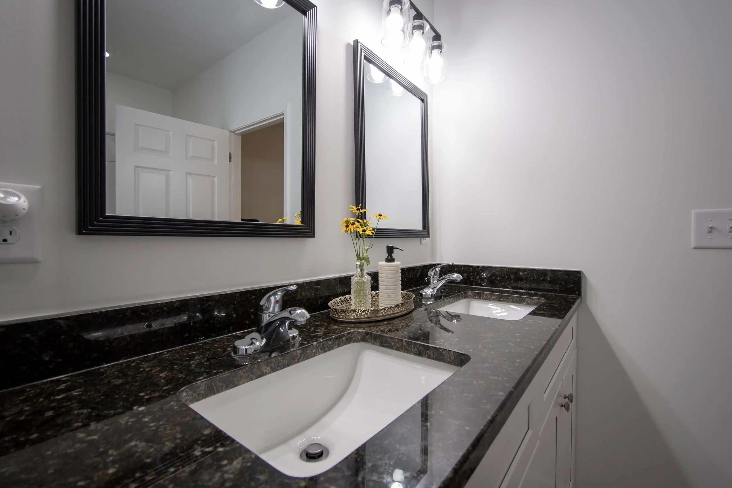 Bathroom with dual sinks, black granite countertop, and two mirrors with black frames. There is a small vase with yellow flowers and a soap dispenser on the counter. The lighting fixture has four bulbs.