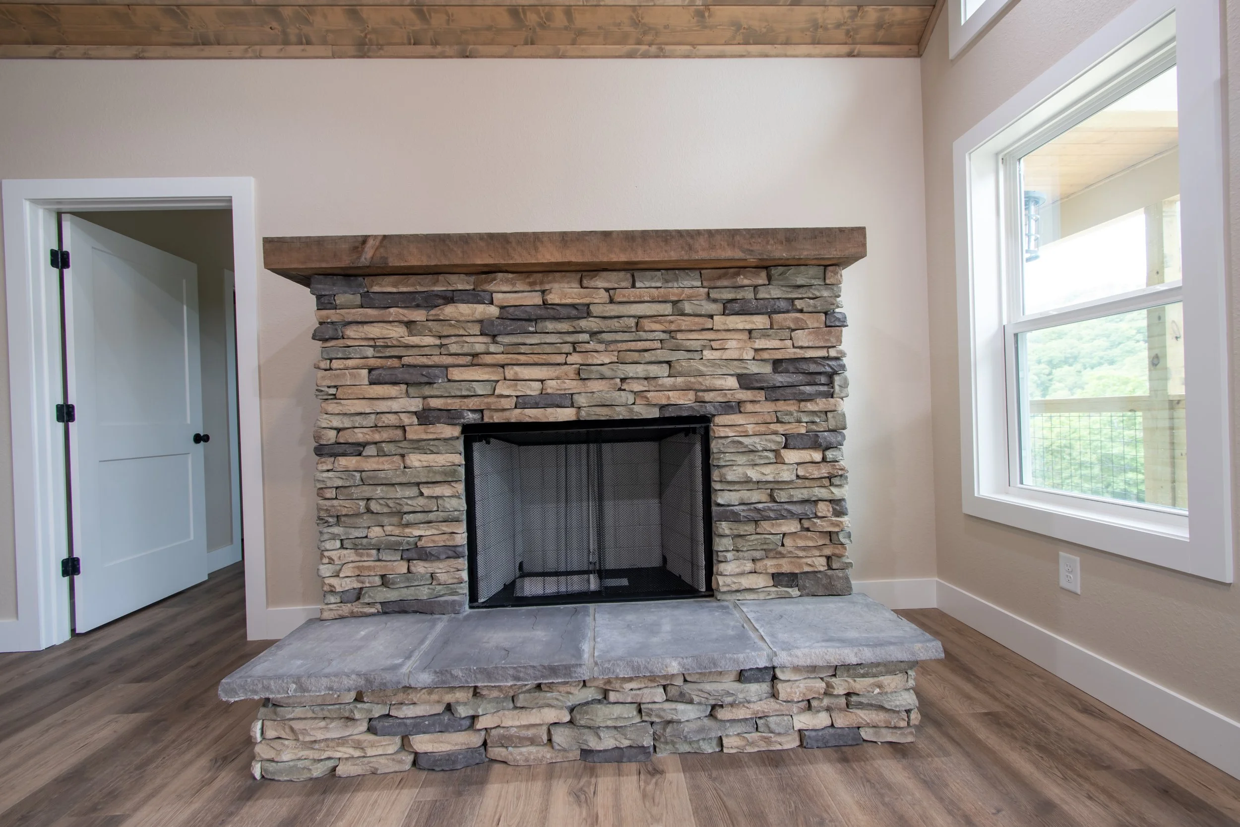 A living room with a stone fireplace, wooden mantel, hardwood floor, open doorway, large window, and white walls.