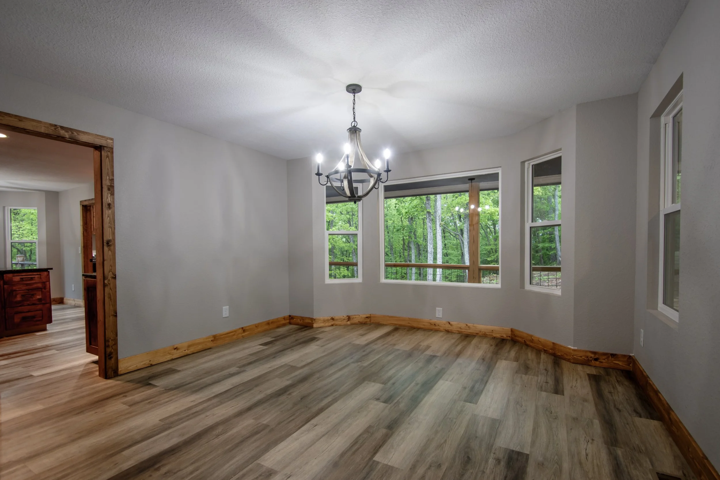 Empty living room with hardwood floors, white walls, multiple windows overlooking a green forest, and a black chandelier hanging from the ceiling.