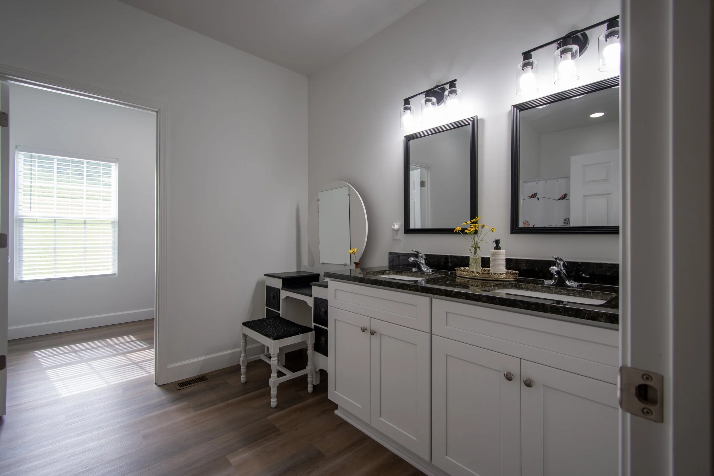 Bathroom with white cabinetry, black granite countertops, two sinks, two mirrors, and modern light fixtures; small makeup vanity with mirror, stool, and decorative tray with a small flower vase and soap dispenser.