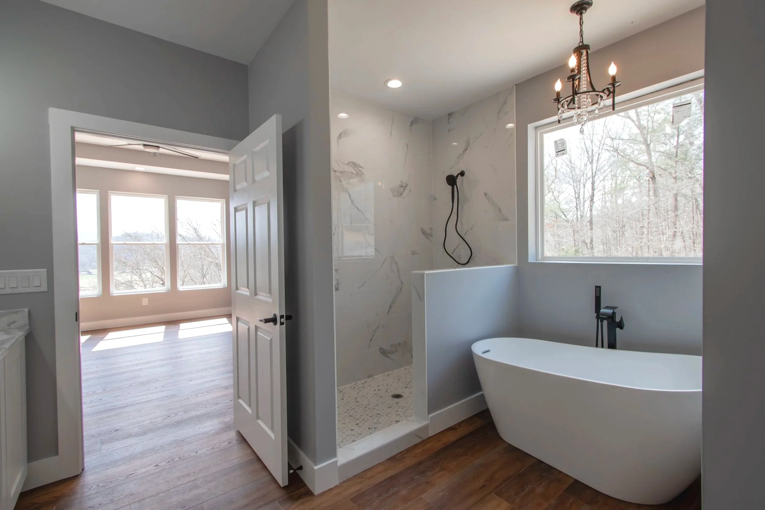 Modern bathroom with a freestanding bathtub, marble shower, large window, chandelier, and hardwood floors