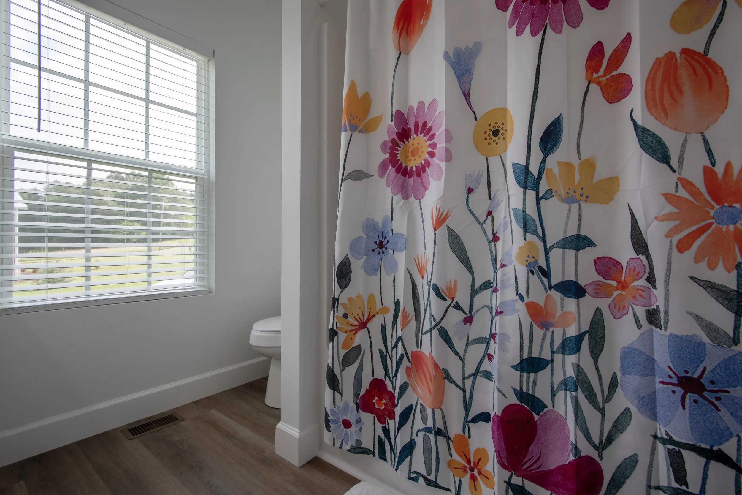 A bathroom with a large window with white blinds, a white toilet, and a shower curtain with colorful floral patterns.