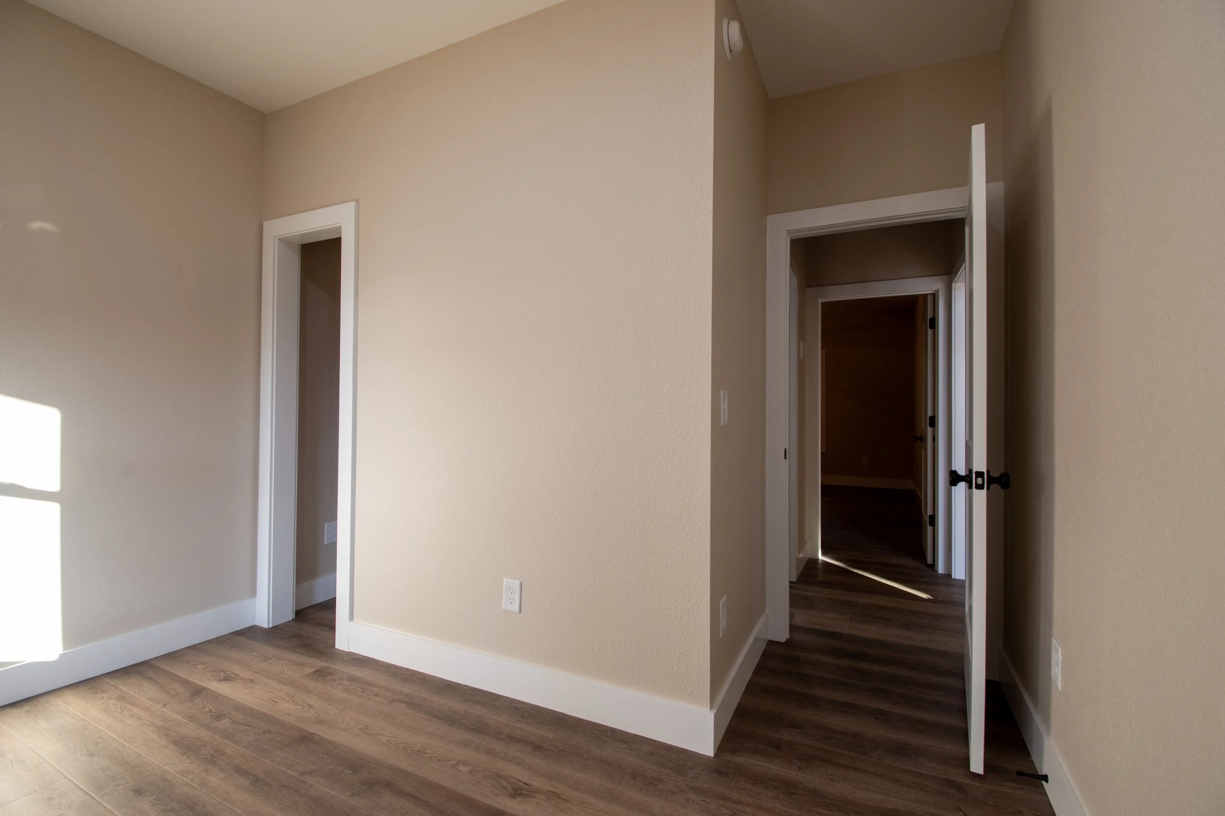 Empty room with beige walls, white baseboards, wood flooring, open doorways leading to other rooms, and sunlight coming through a window.