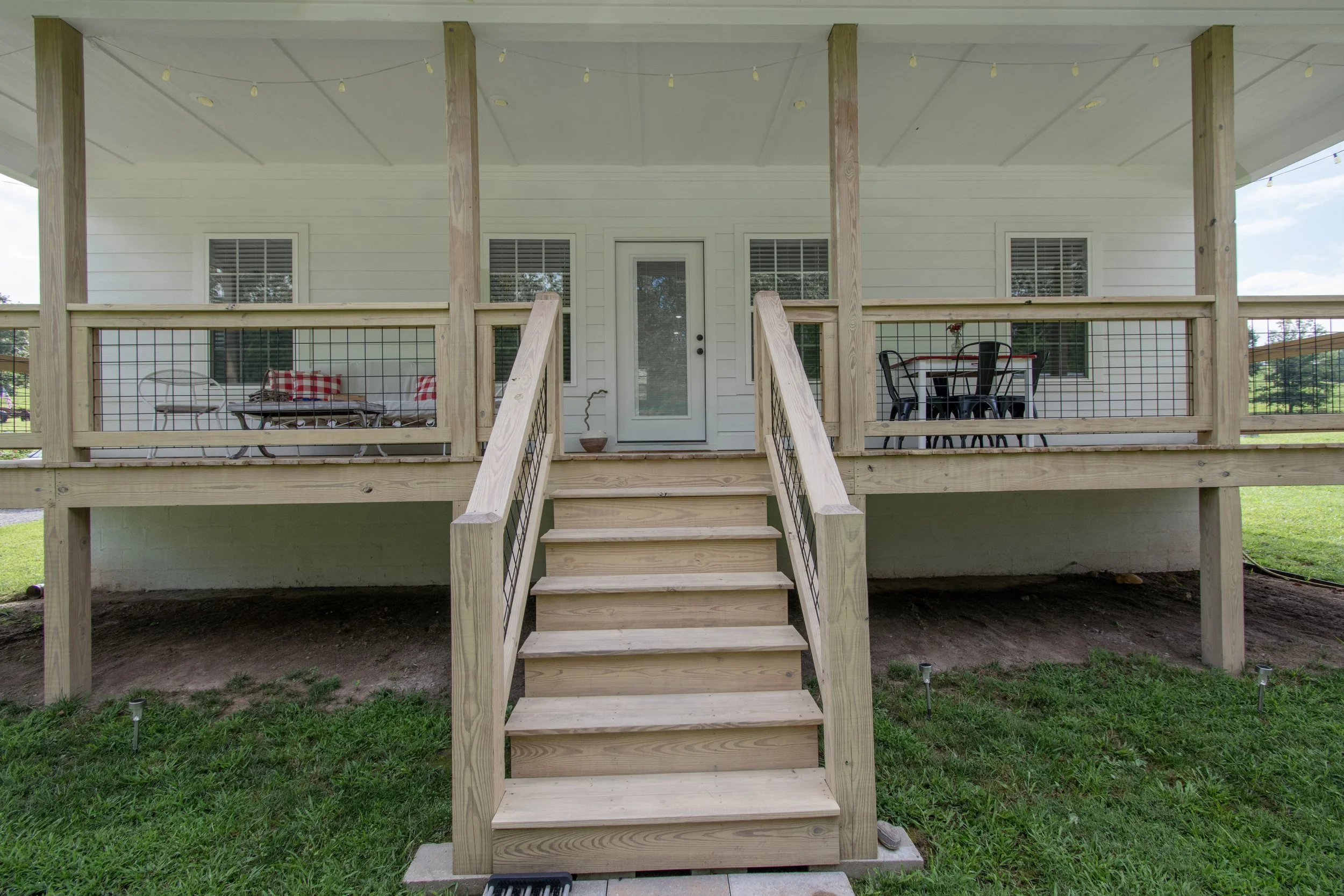 Newly constructed wooden deck with stairs leading up to a white house with a screened door and windows, with outdoor seating on the deck in a backyard.