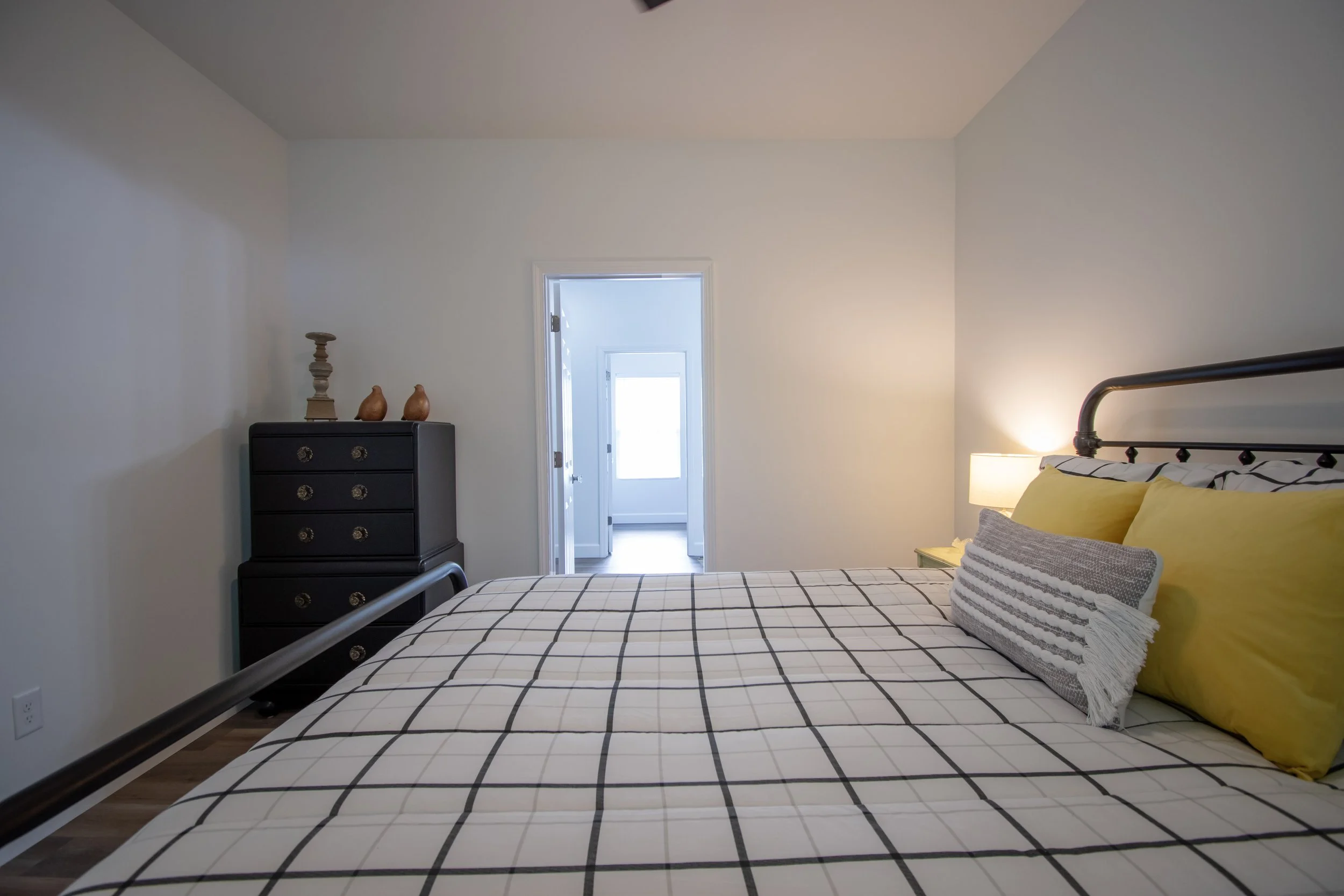 Bedroom with bed, black metal headboard, yellow pillows, black and white grid bedding, nightstand with lamp, black chest of drawers with decorative items, doorway leading to a bright windowed room.