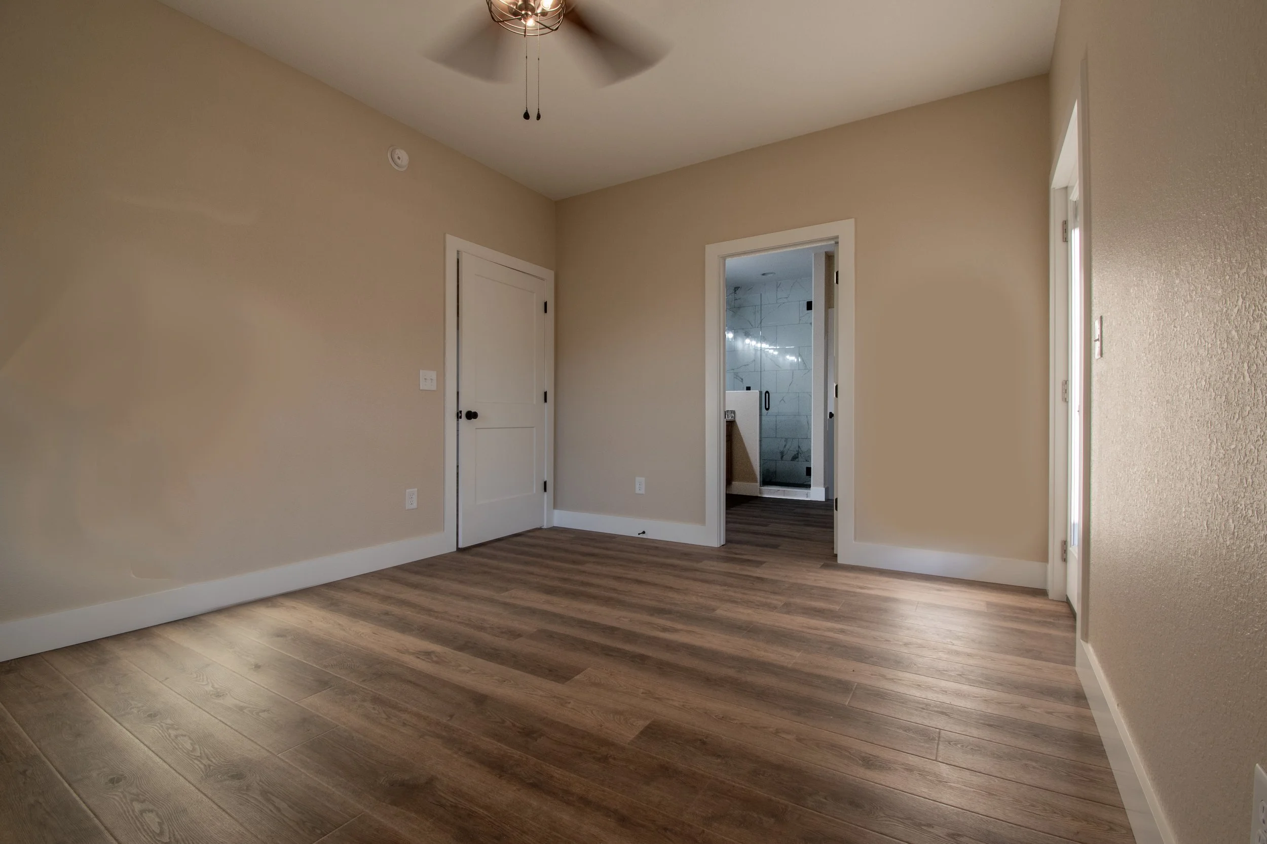 Empty room with wood flooring, beige walls, a ceiling fan, and a door leading to a bathroom with white and gray tile.