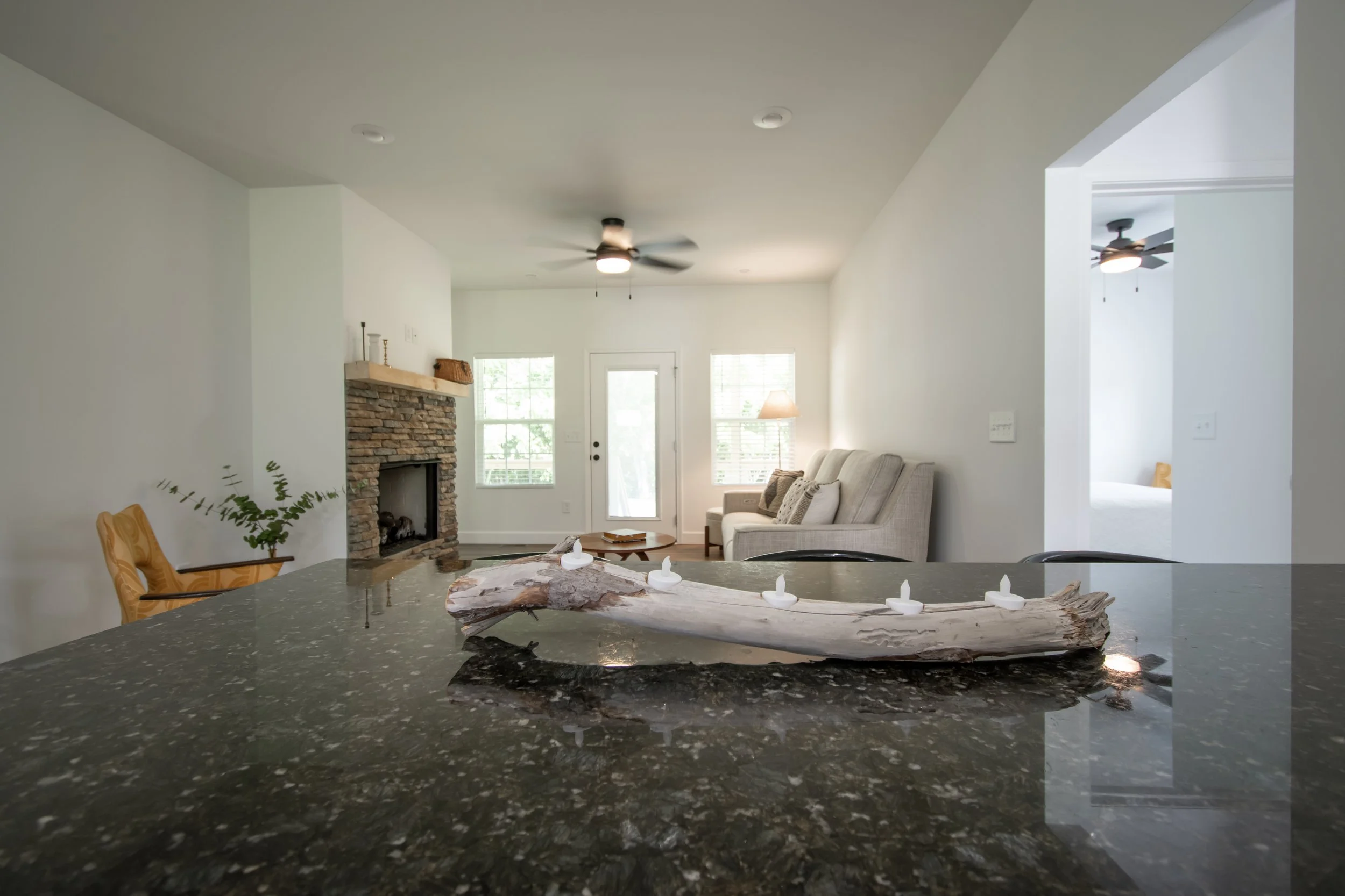 Open concept living room with a fireplace, beige upholstered sofa, ceiling fans, and natural light from windows, viewed from a kitchen countertop with a decorative driftwood and candle centerpiece.