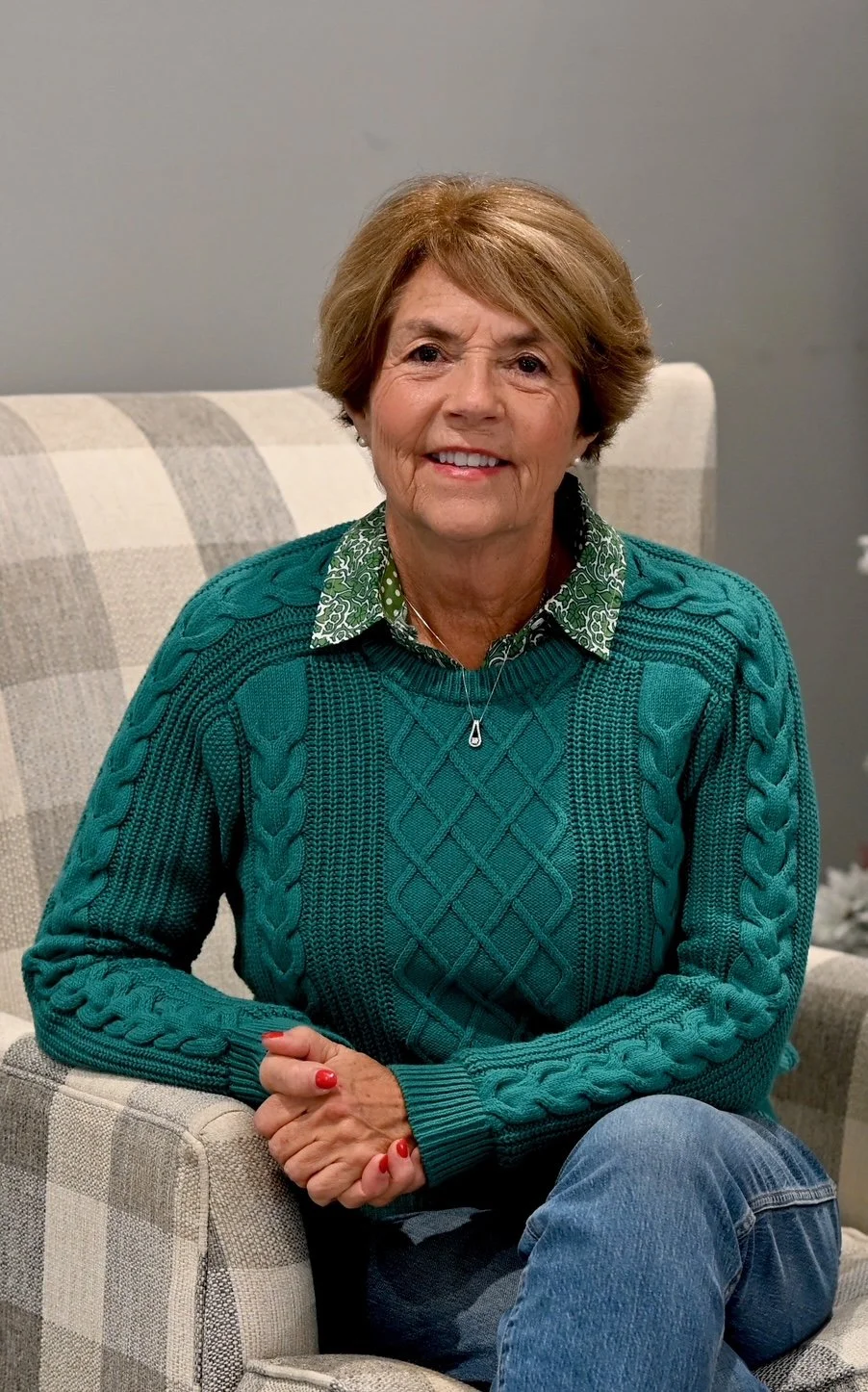 A smiling elderly woman with short light brown hair, wearing a green cable-knit sweater over a patterned green and white blouse, sitting on a beige plaid armchair against a gray wall.