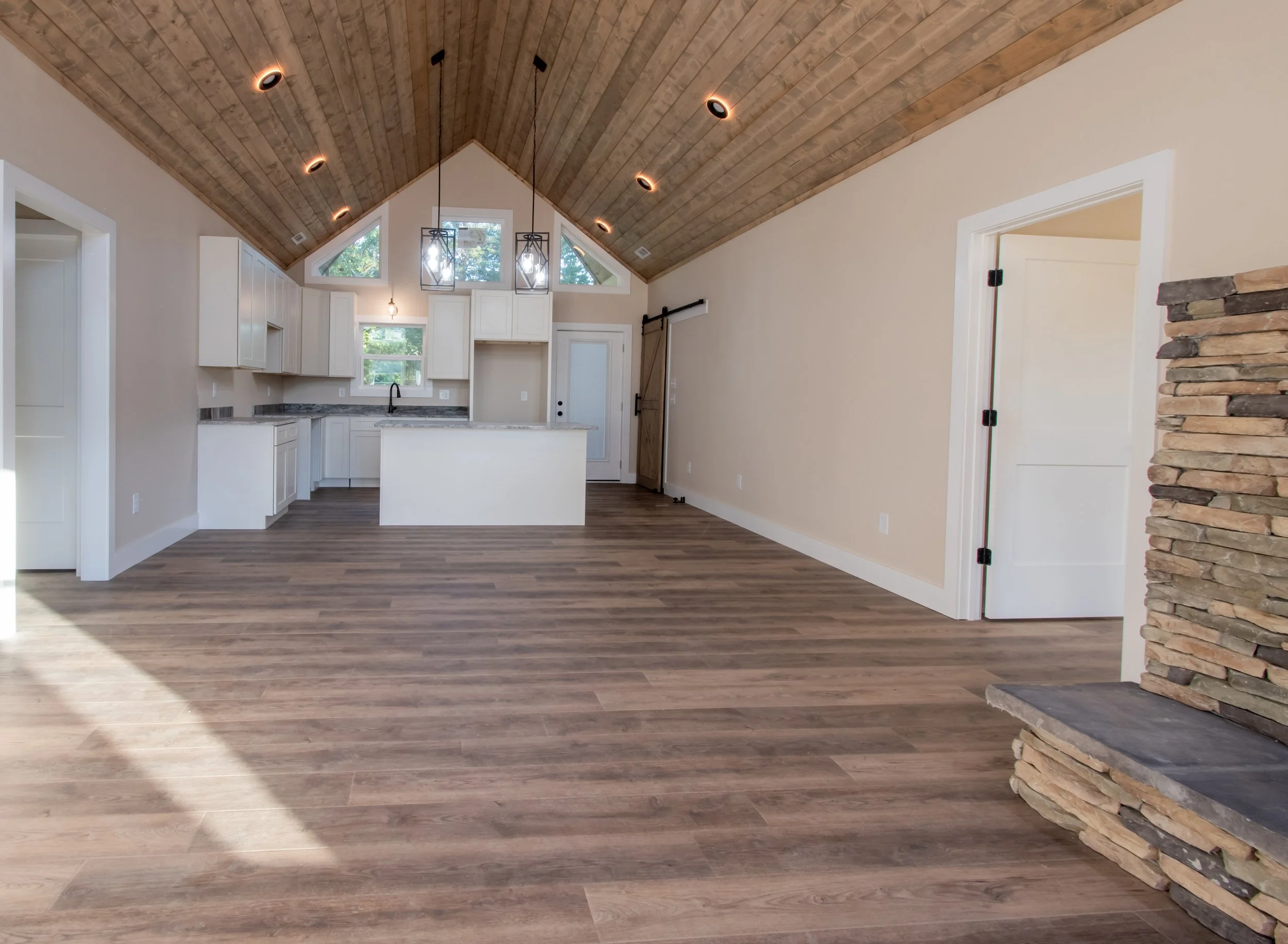 Empty open-concept living room and kitchen with wooden vaulted ceiling, white cabinets, stone fireplace, and hardwood flooring.