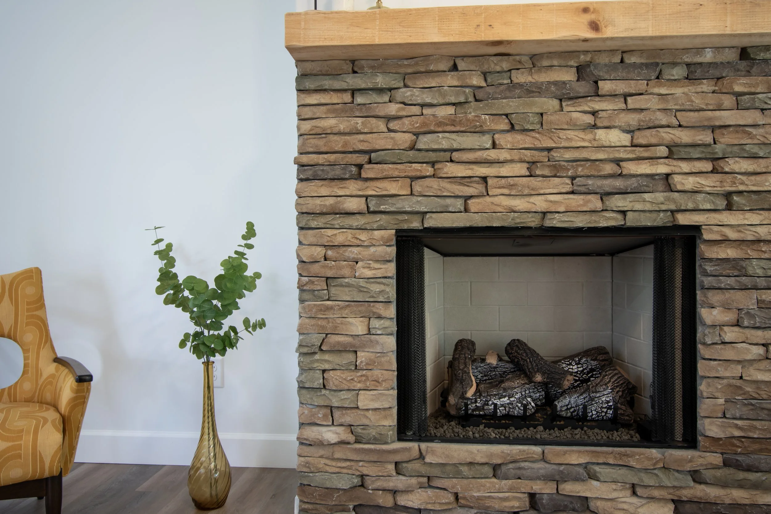 A stone fireplace with logs inside, next to a yellow armchair and a tall vase with green leafy plant, in a room with white walls and wood flooring.