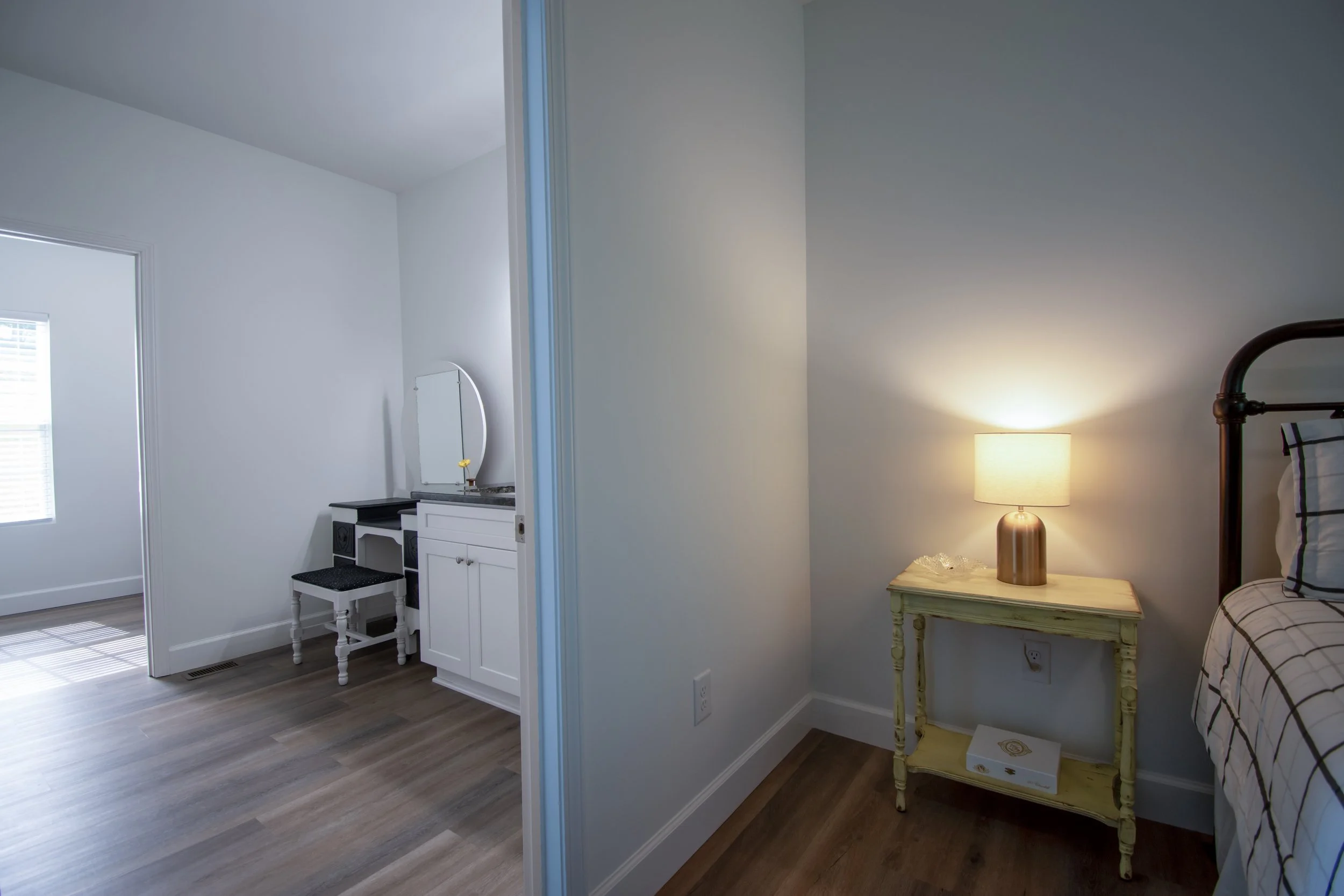 A view of a bedroom with a nightstand and lamp on the right, and part of an adjacent room with a vanity and stool on the left, showing wood flooring and white walls.