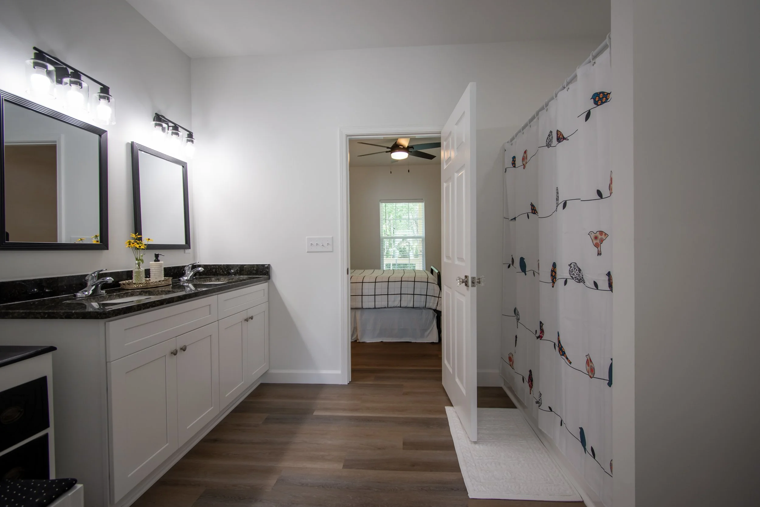Bathroom with double sinks, black granite countertop, white cabinets, and two mirrors. A vase with yellow flowers, soap dispenser, and decorative tray are on the countertop. Light fixtures above the mirrors. To the right, a white closet door with a c