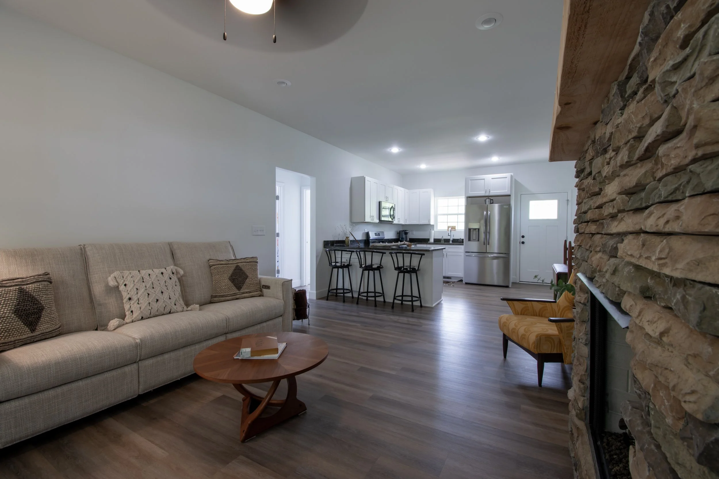 Living room with beige couch, wooden coffee table, stone fireplace, and kitchen with white cabinets, stainless steel appliances, and bar stools.