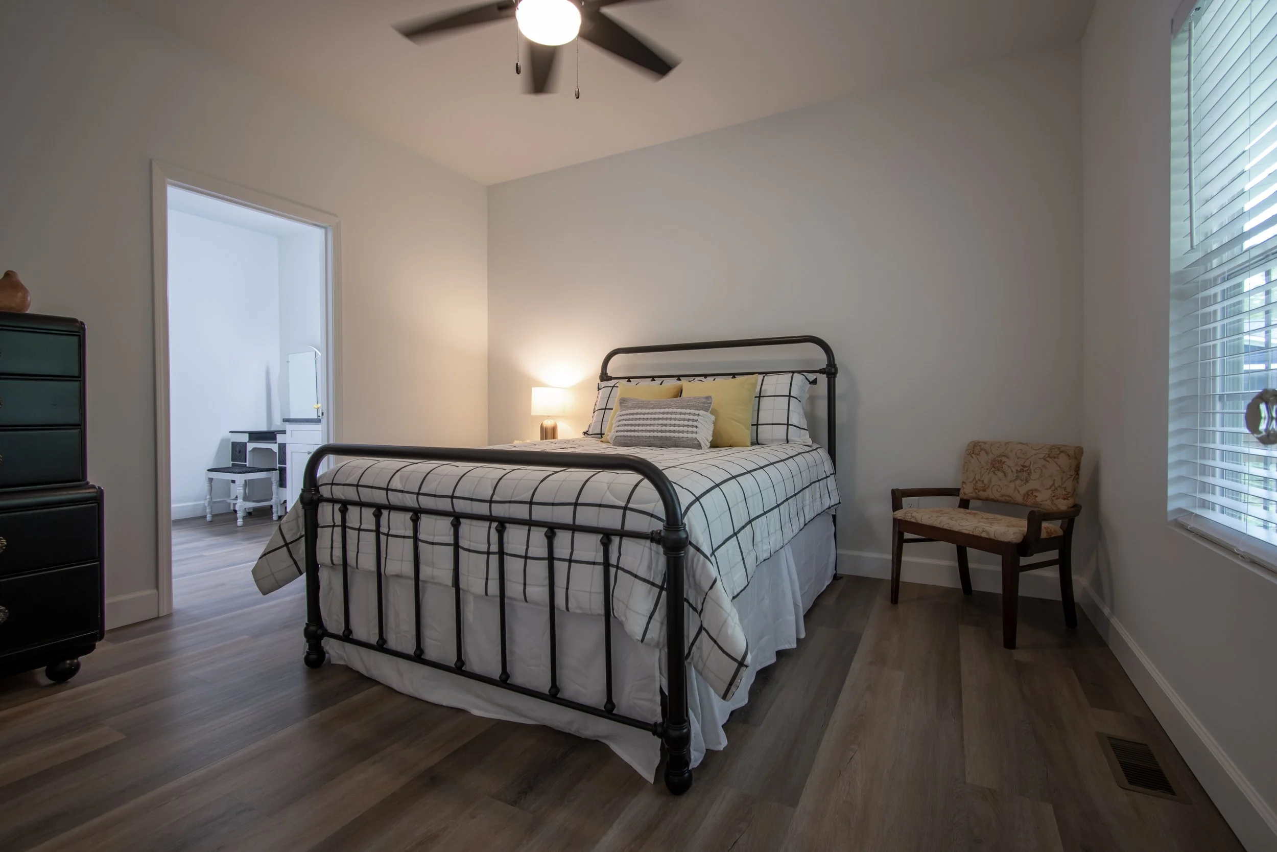 A bedroom with a black metal bed frame, checkered bedding, and multiple pillows in front of a white wall. There is a small wooden chair with floral upholstery next to a large window with blinds. A door leads to another room, and a ceiling fan is on t