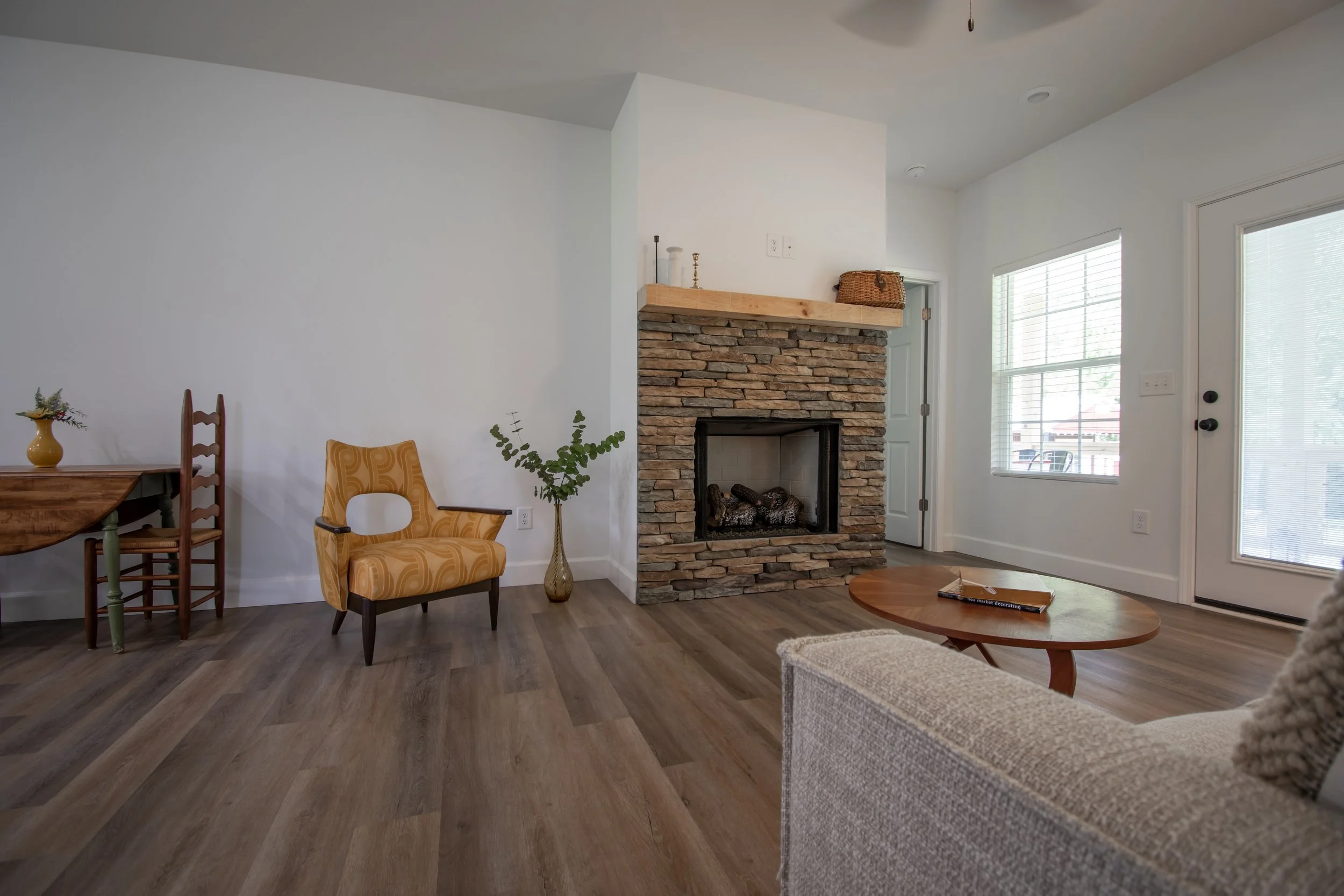 Living room with fireplace, yellow patterned armchair, wooden coffee table, gray sofa, wooden dining table, and large windows with blinds.