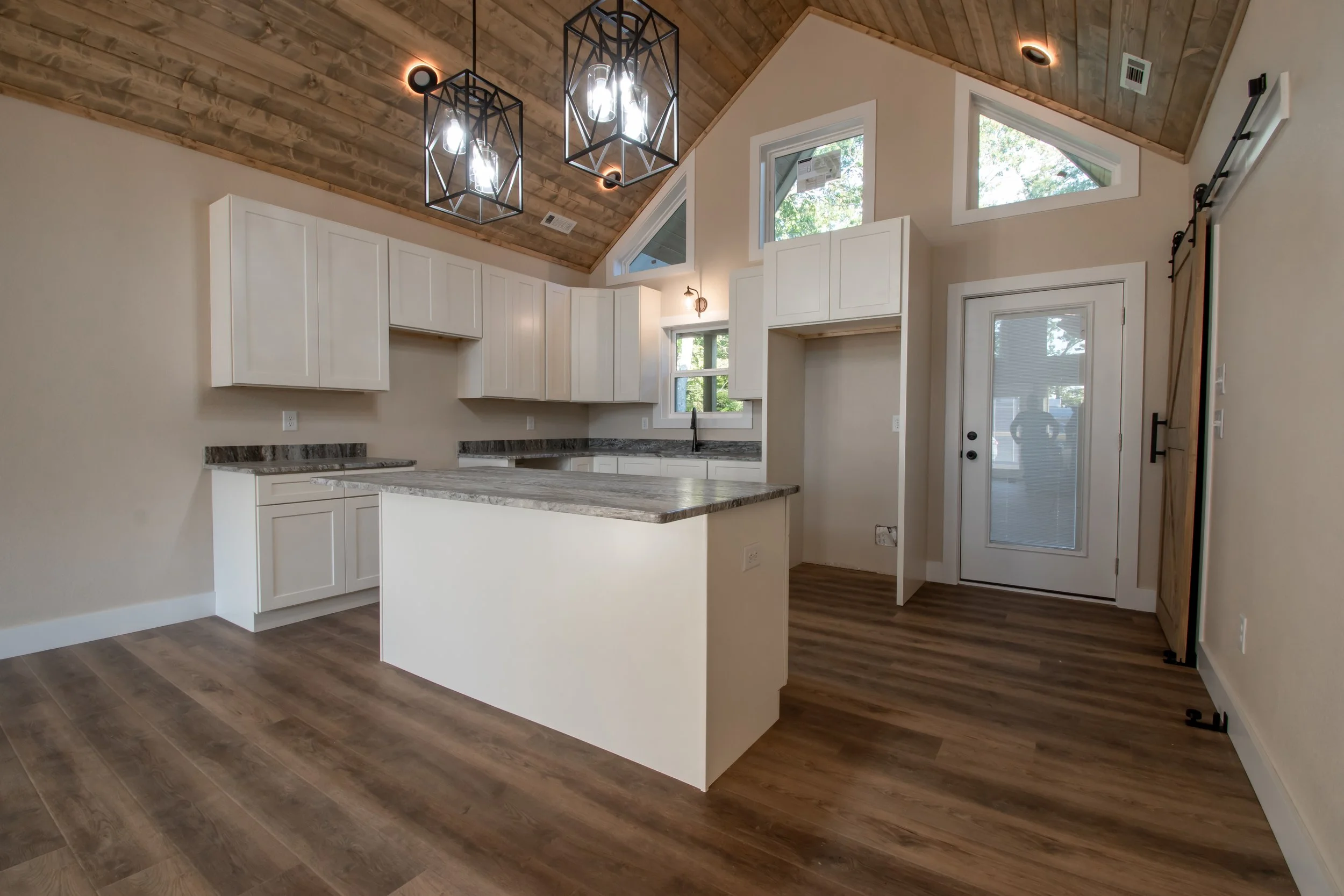 Interior view of a modern kitchen with white cabinets, a large island with a stone countertop, wood flooring, and a vaulted wooden ceiling with hanging light fixtures. There are windows and a door leading outside.
