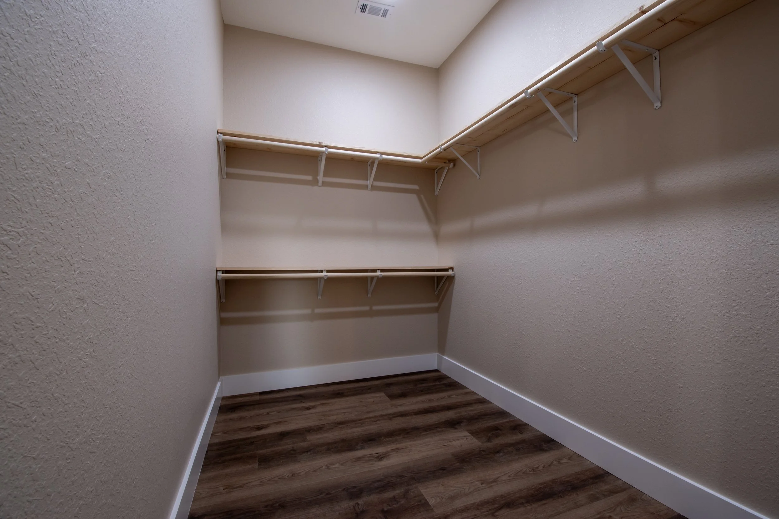 Empty walk-in closet with beige walls, wooden shelves, and a dark wood floor.
