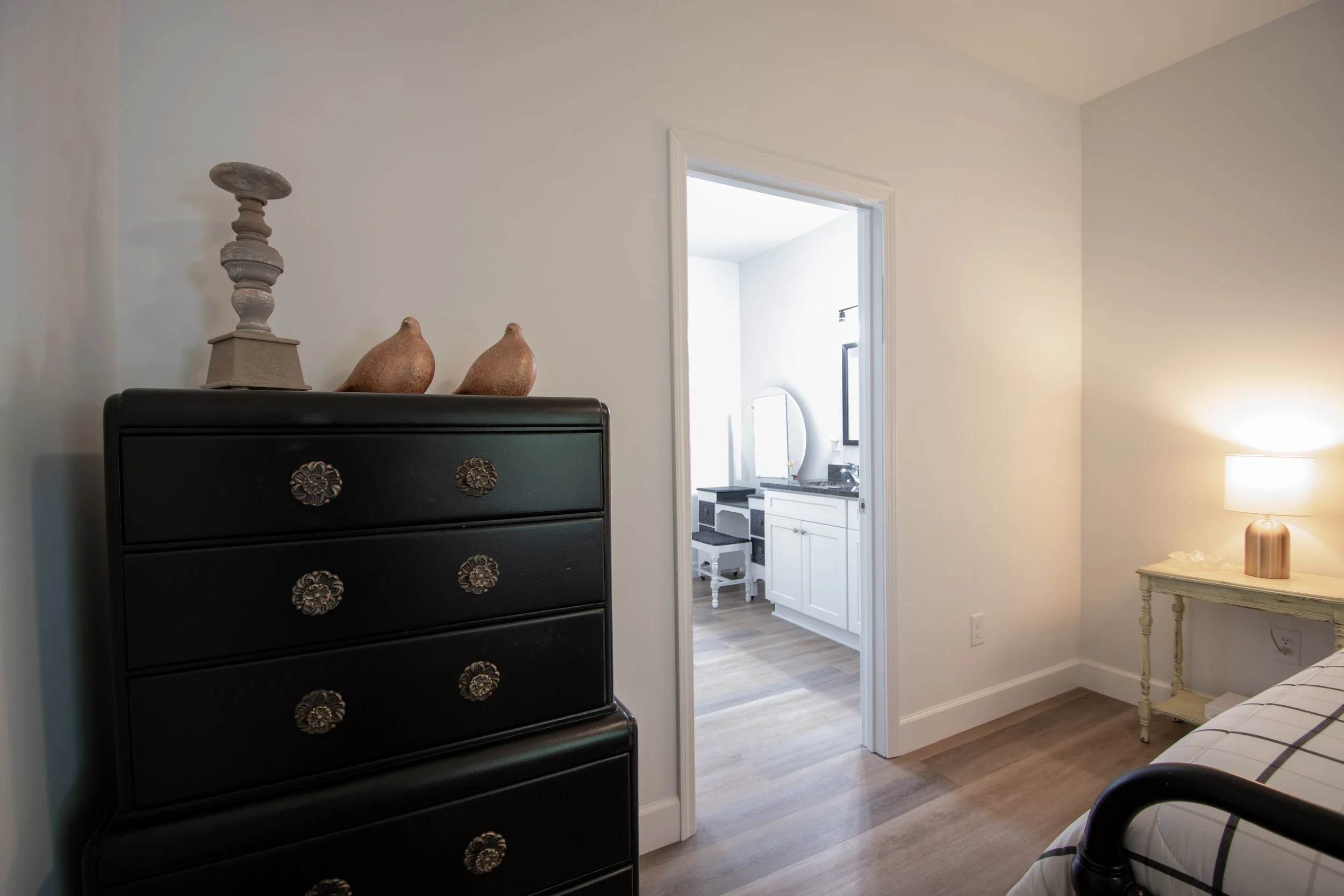 View of a bedroom with a black dresser, decorative objects on top, and a nightstand with a lamp. An open door reveals a bathroom with white cabinetry and a mirror.