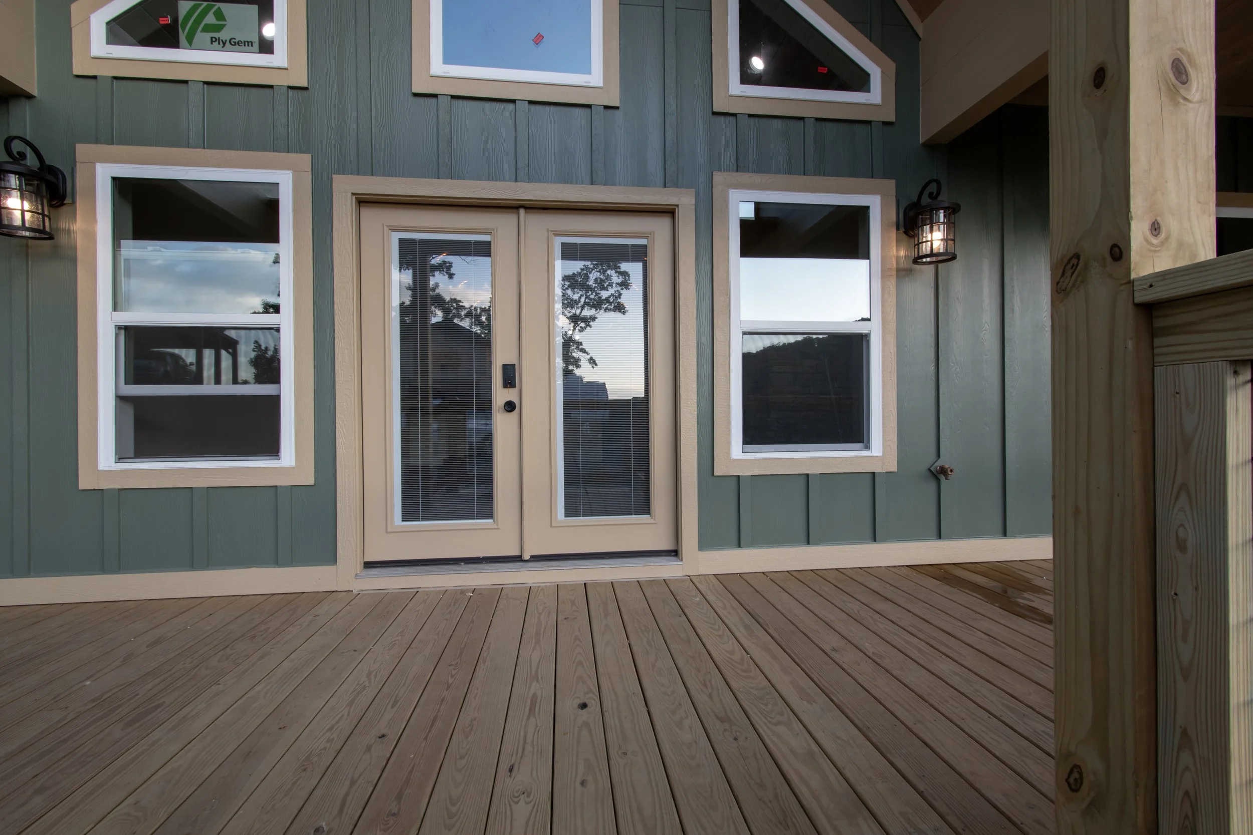 Image of a house exterior with green siding, multiple windows, and a French door opening onto a wooden deck, with a mountain and sky reflection in the windows.