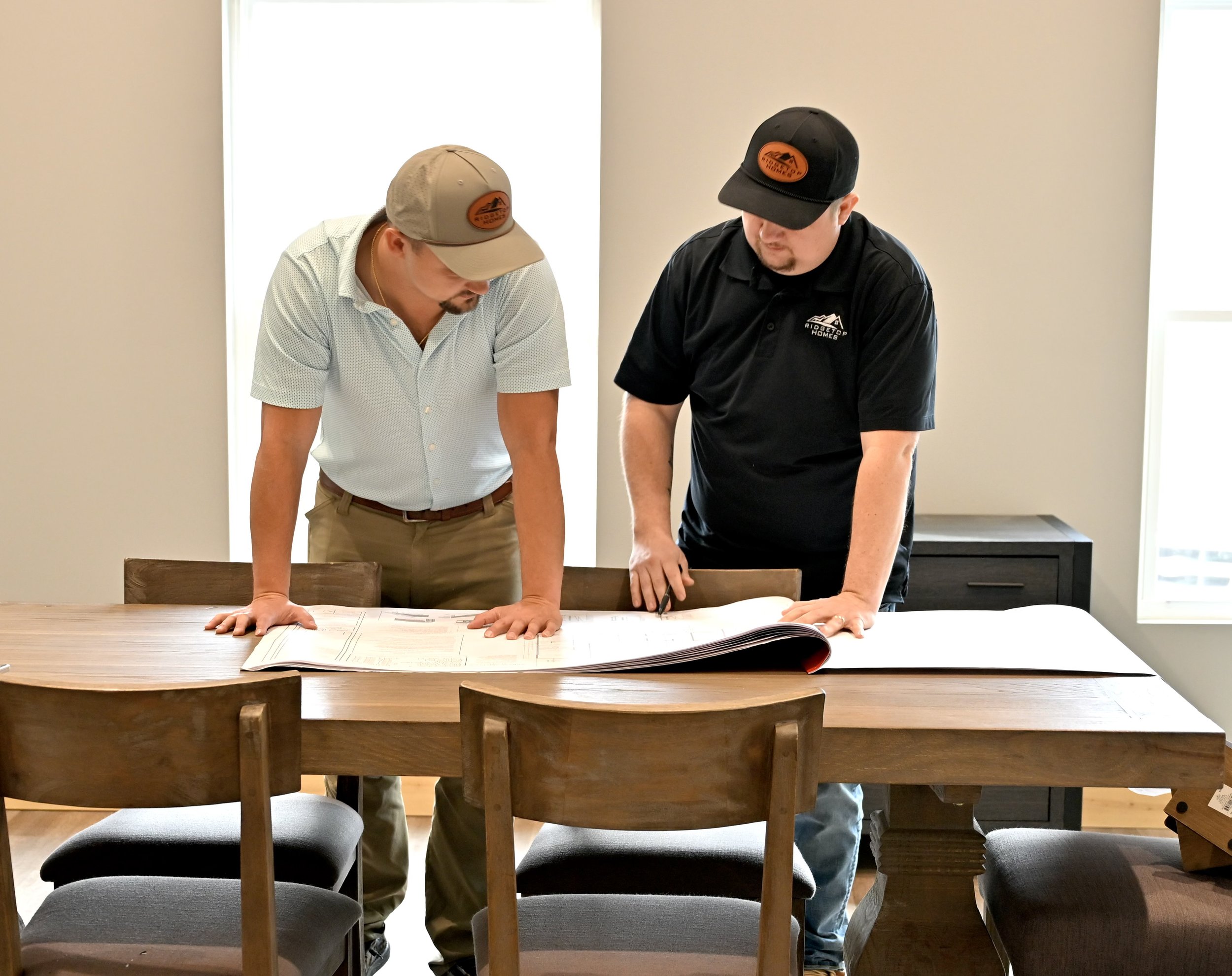 Two men wearing hats and casual shirts are reviewing large documents spread out on a wooden table in a bright room.