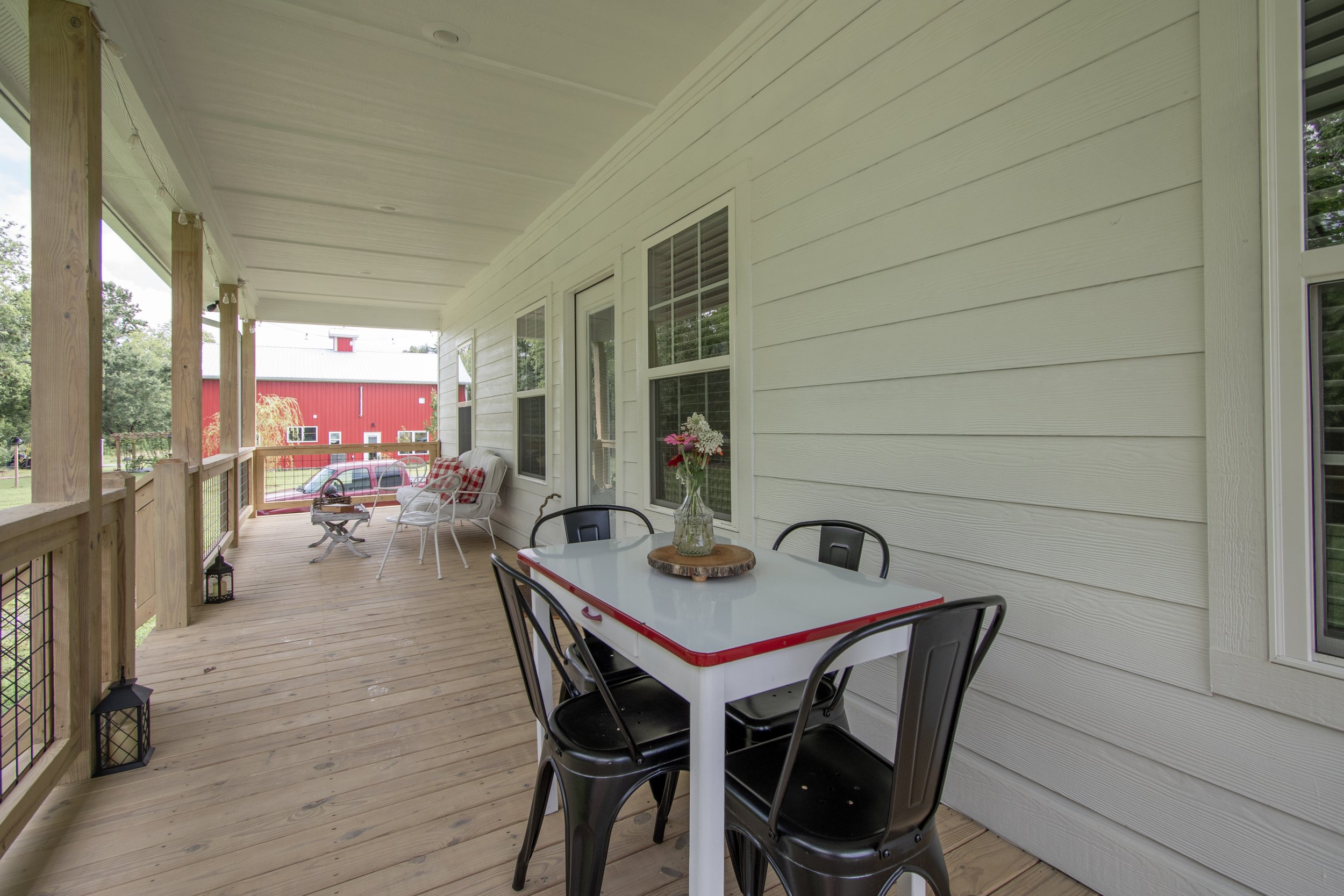 Part of a porch with a white table, black metal chairs, and a clear vase with pink, white, and purple flowers on a wooden slice. There are additional white chairs with red plaid pillows, a small table, lanterns on the floor, and a red barn in the bac