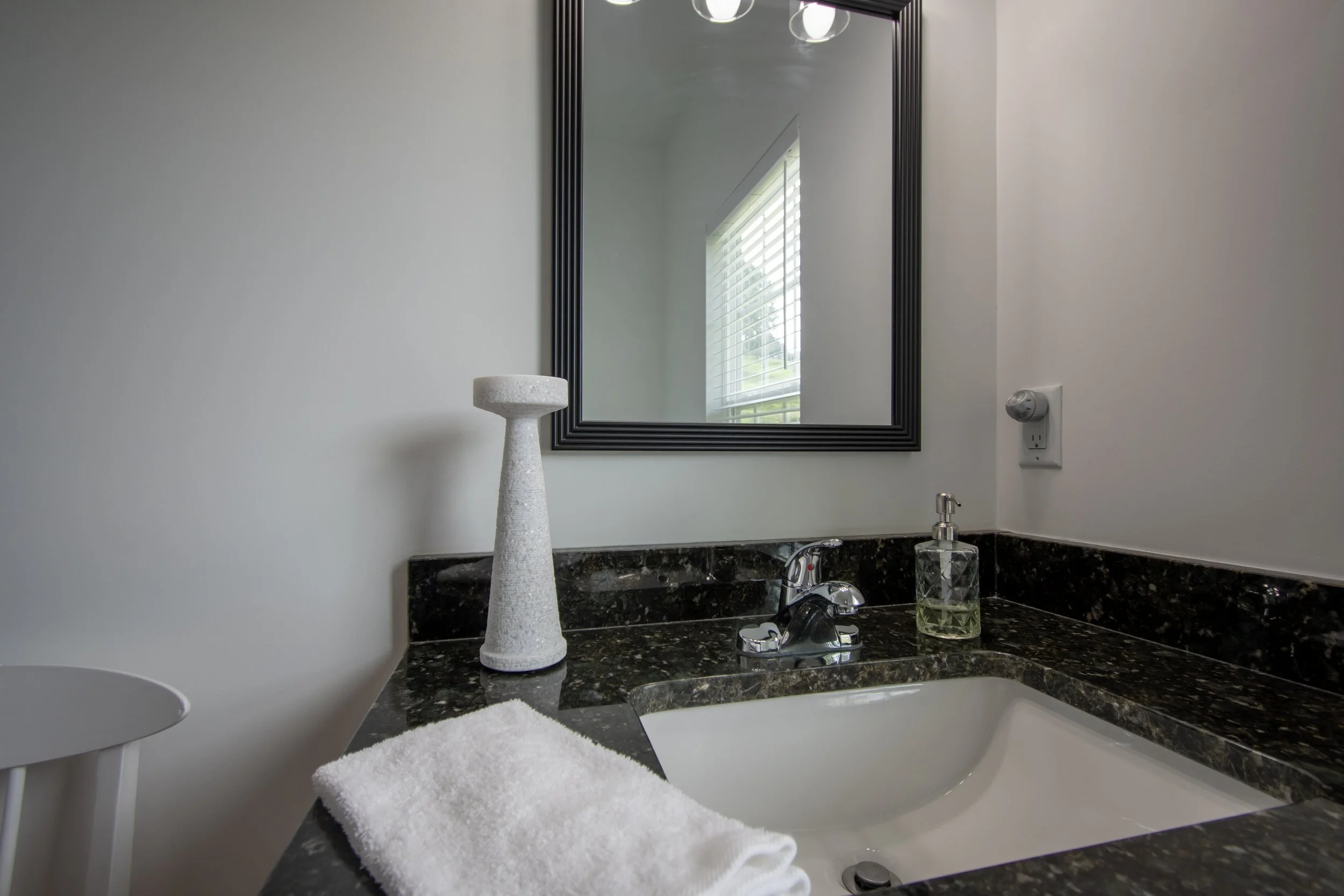 Bathroom vanity with black granite countertop, white sink, mirror with black frame, soap dispenser, decorative container, and a white towel.
