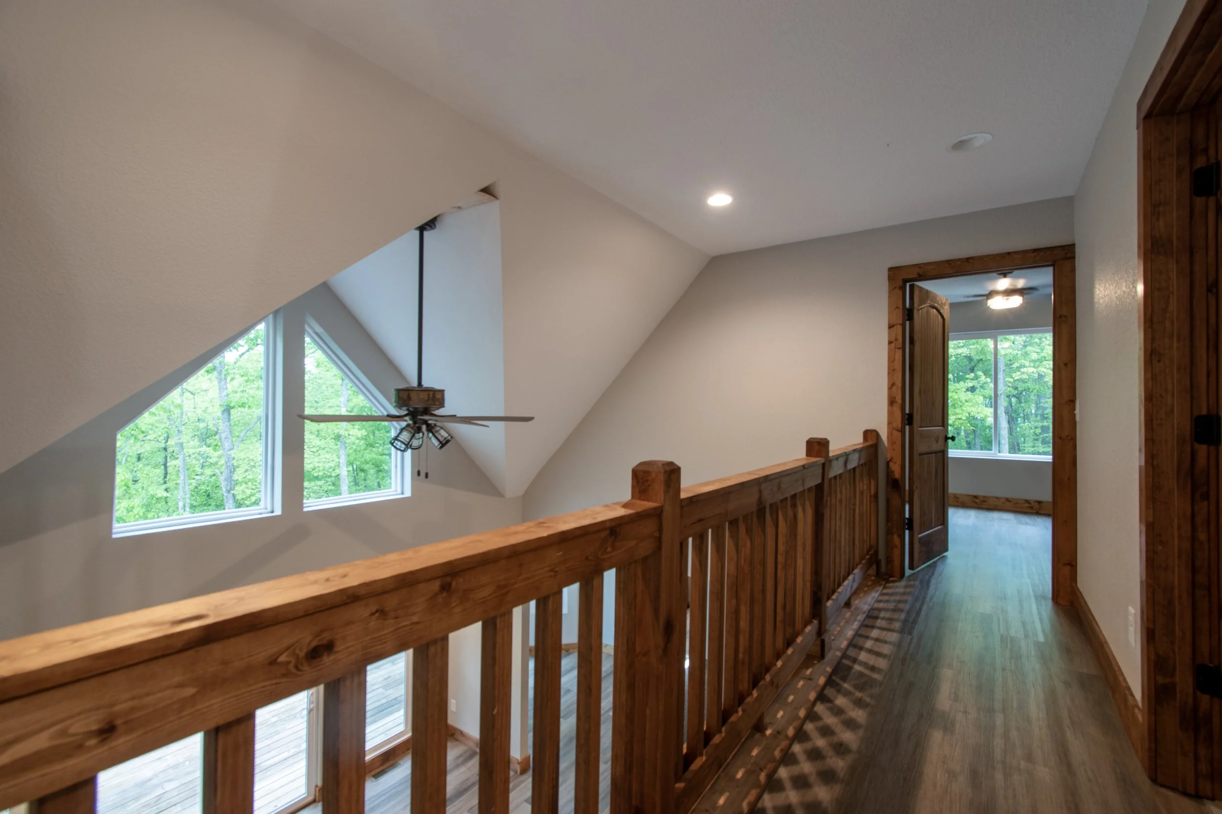 Interior view of a house hallway with wooden railing, angled ceiling with a ceiling fan, and large windows showing green trees outside.