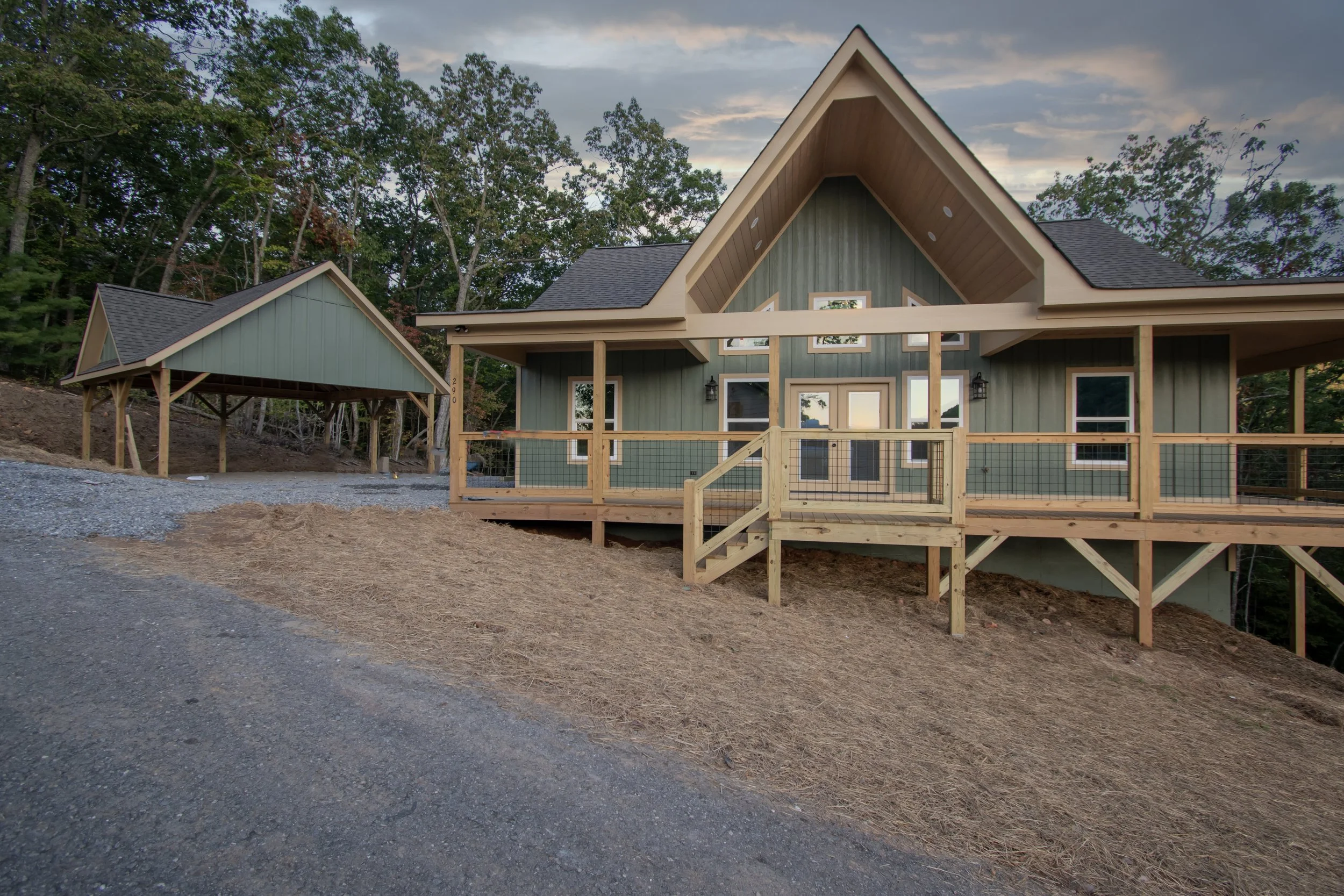 New house with green siding and a large front porch under construction, surrounded by trees and a gravel driveway.