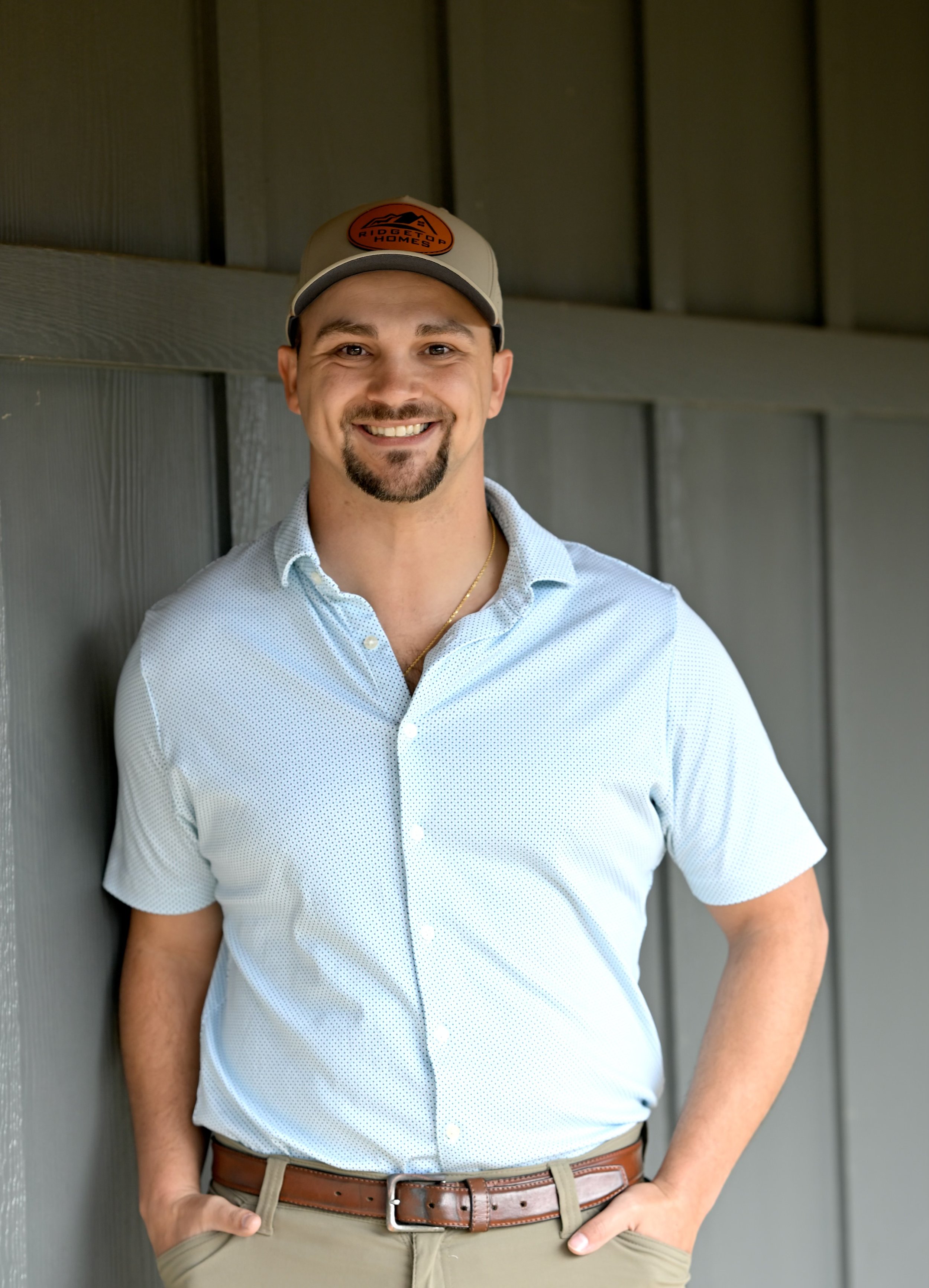 A smiling man with a beard and mustache wearing a beige cap, a light blue button-up shirt, and khaki pants, standing against a wooden wall.