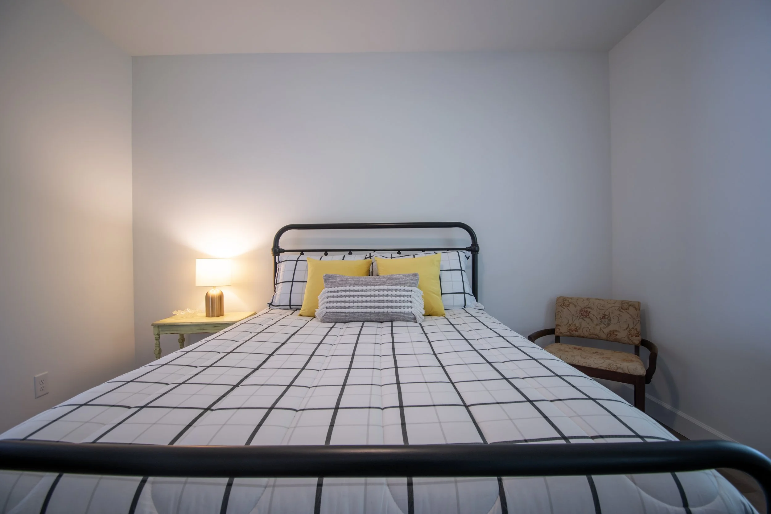 A neatly made bed with a black metal frame, decorated with yellow, gray, and striped pillows, is situated next to a lit bedside lamp on a small table in a minimalist bedroom with light-colored walls.