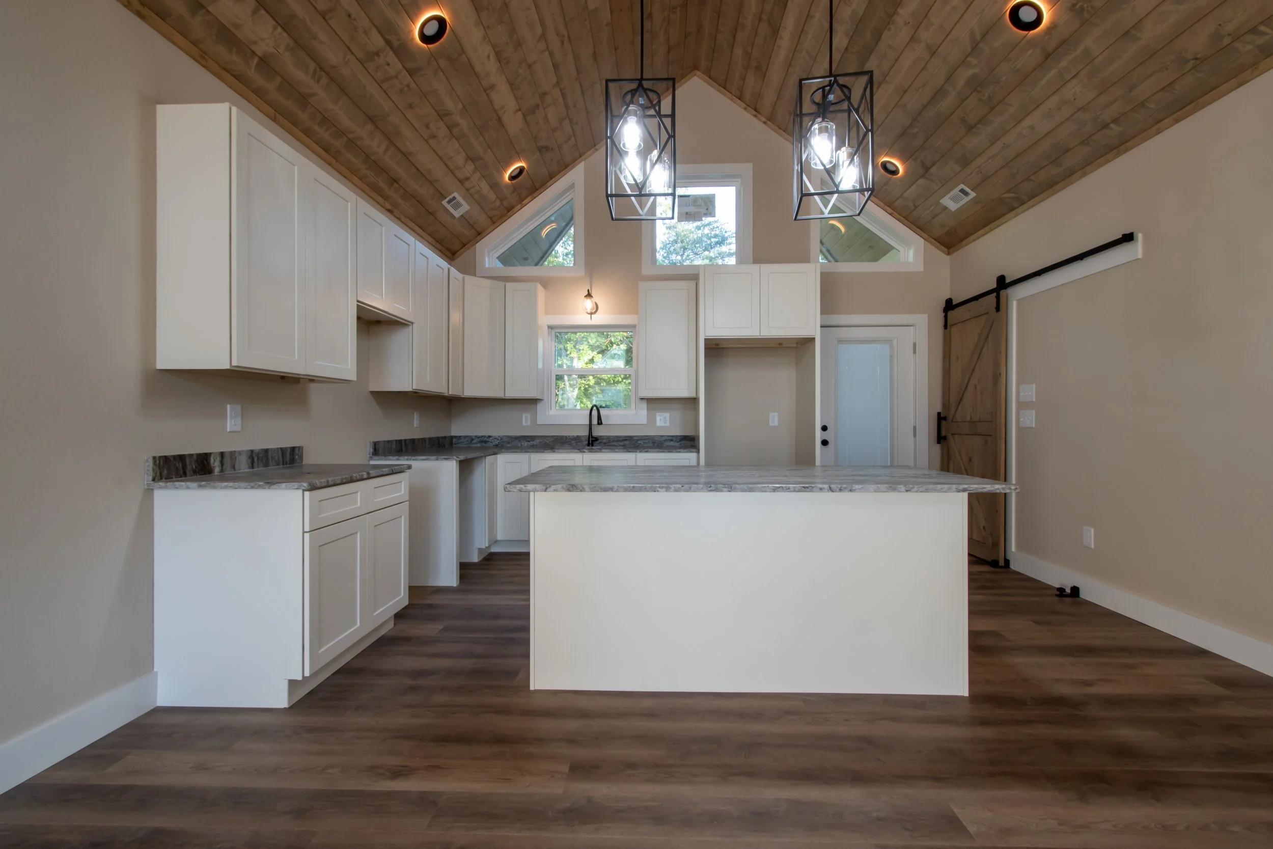 Modern kitchen with white cabinets, marble countertops, and wooden ceiling, featuring pendant lighting and barn sliding door.