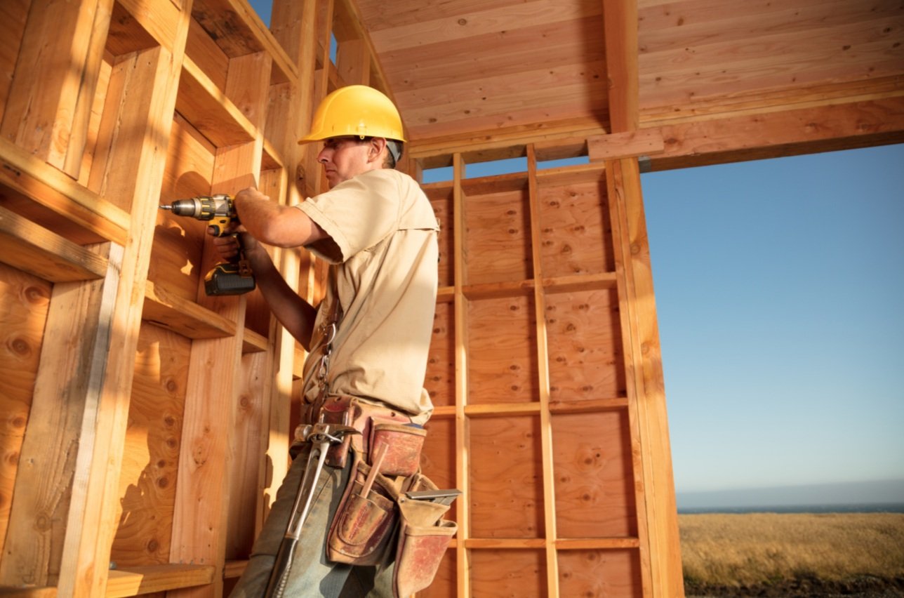 A construction worker wearing a yellow hard hat and beige shirt uses a cordless drill on a wooden wall frame inside a partially built structure, with an open wall showing an outdoor landscape and clear sky.