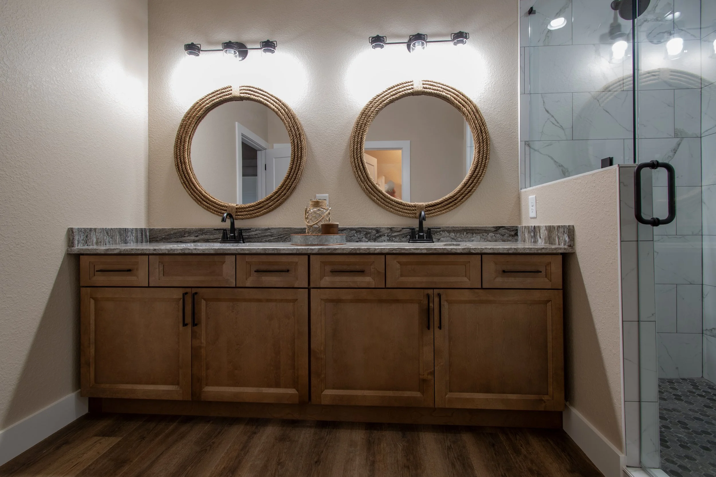 Bathroom vanity with two round woven mirrors, two black faucets, a granite countertop, wooden cabinets, decorative items on tray, and a glass shower with marble tiles.