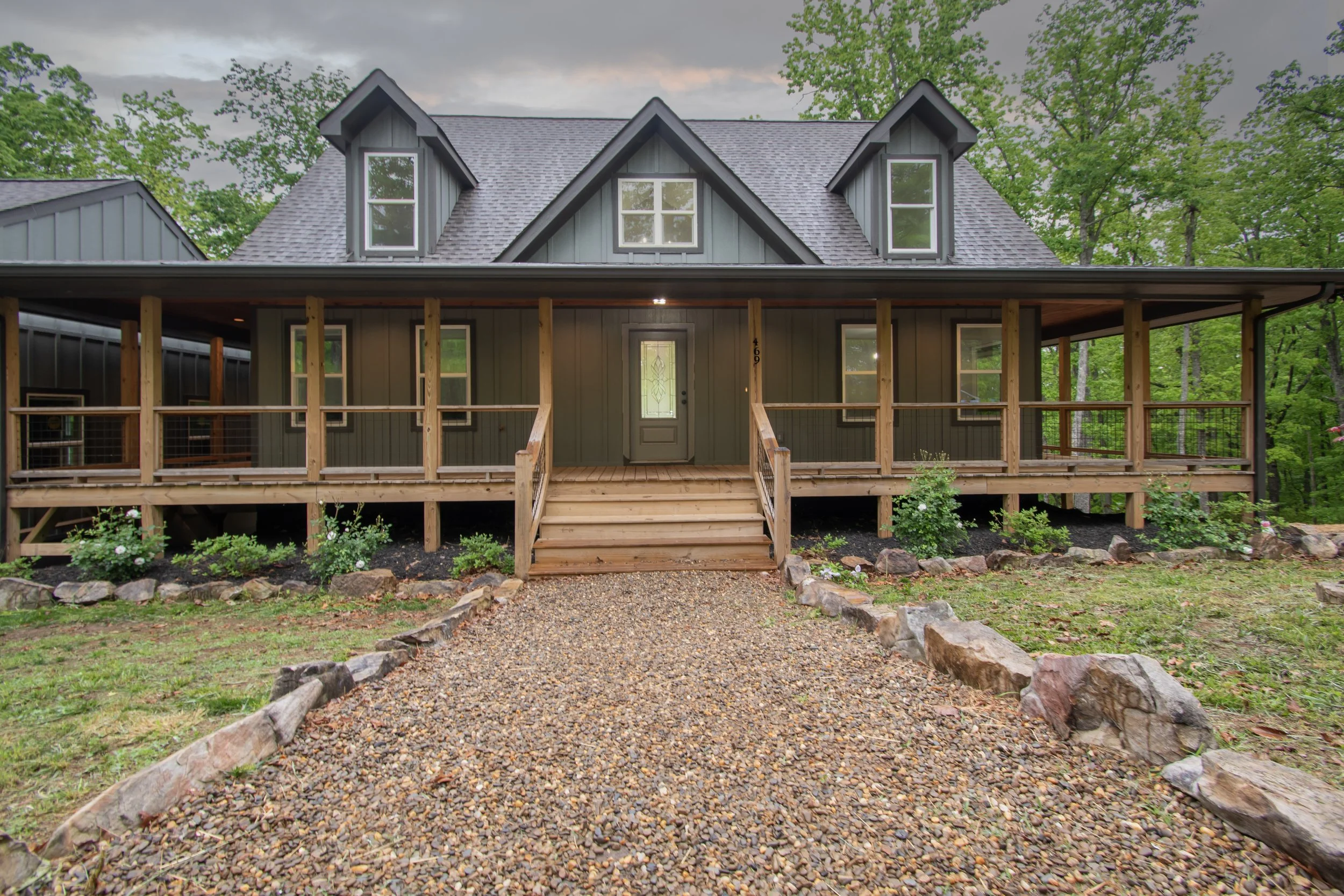 Front view of a house with a wooden porch and stairs, set in a wooded area, with trees and shrubs surrounding it.