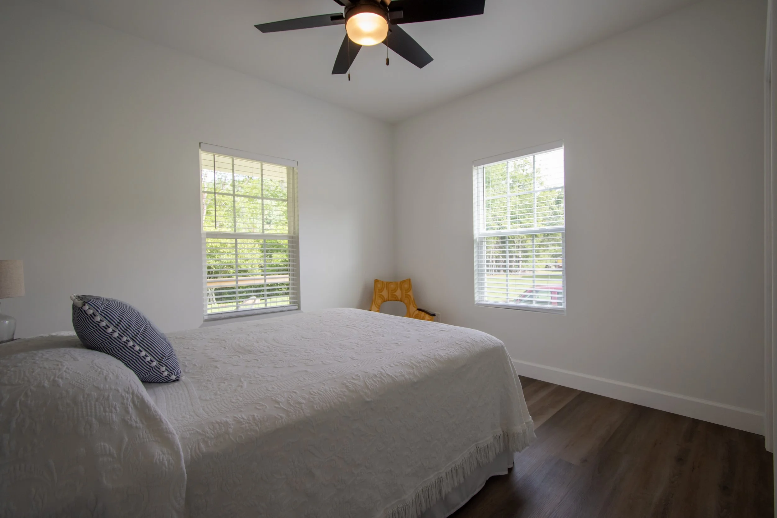 A bedroom with white walls, a ceiling fan with a light, a bed with a white quilt and striped pillow, a window with blinds showing greenery outside, and a yellow chair in the corner.