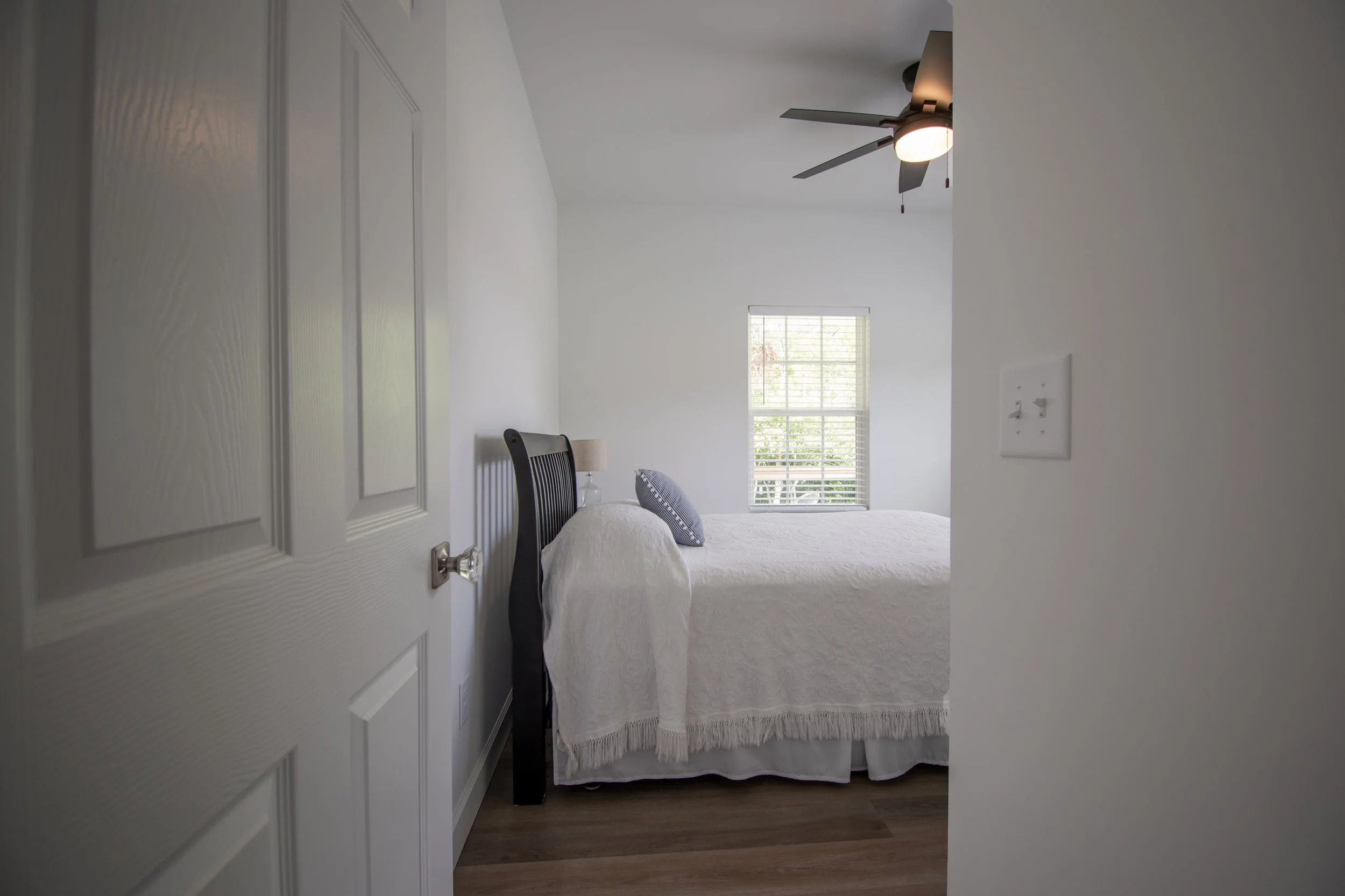 A bedroom with white walls, a window with blinds letting in natural light, a bed with a white quilt and pillows, a wooden headboard, a small nightstand with a lamp, a ceiling fan, and a light switch on the wall.