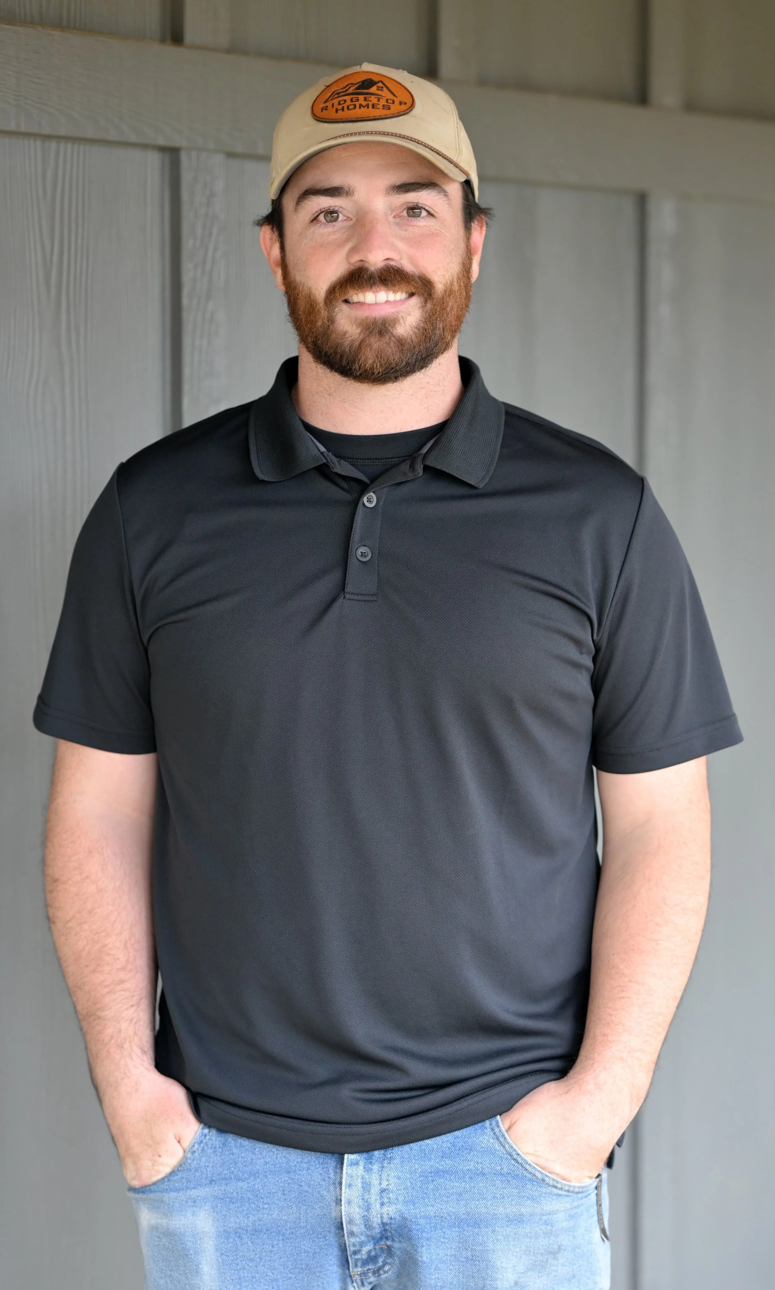 A man with a beard wearing a beige hat with an orange logo, a black polo shirt, and blue jeans, standing in front of a gray wooden wall.