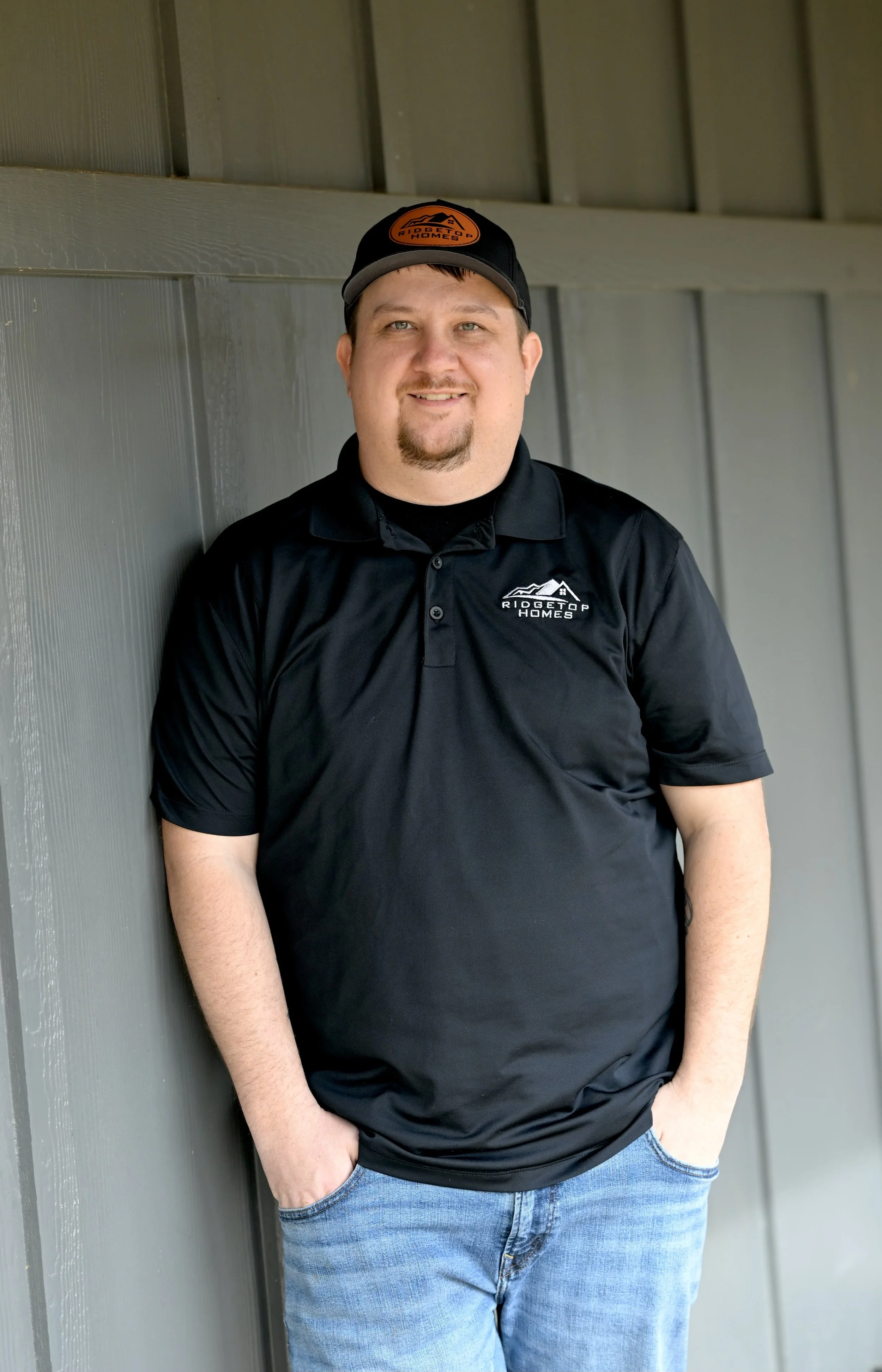 A man with short hair and a goatee, wearing a black polo shirt and a black cap, standing against a gray wall. The shirt and cap both display the 'Ridgetop Homes' logo.