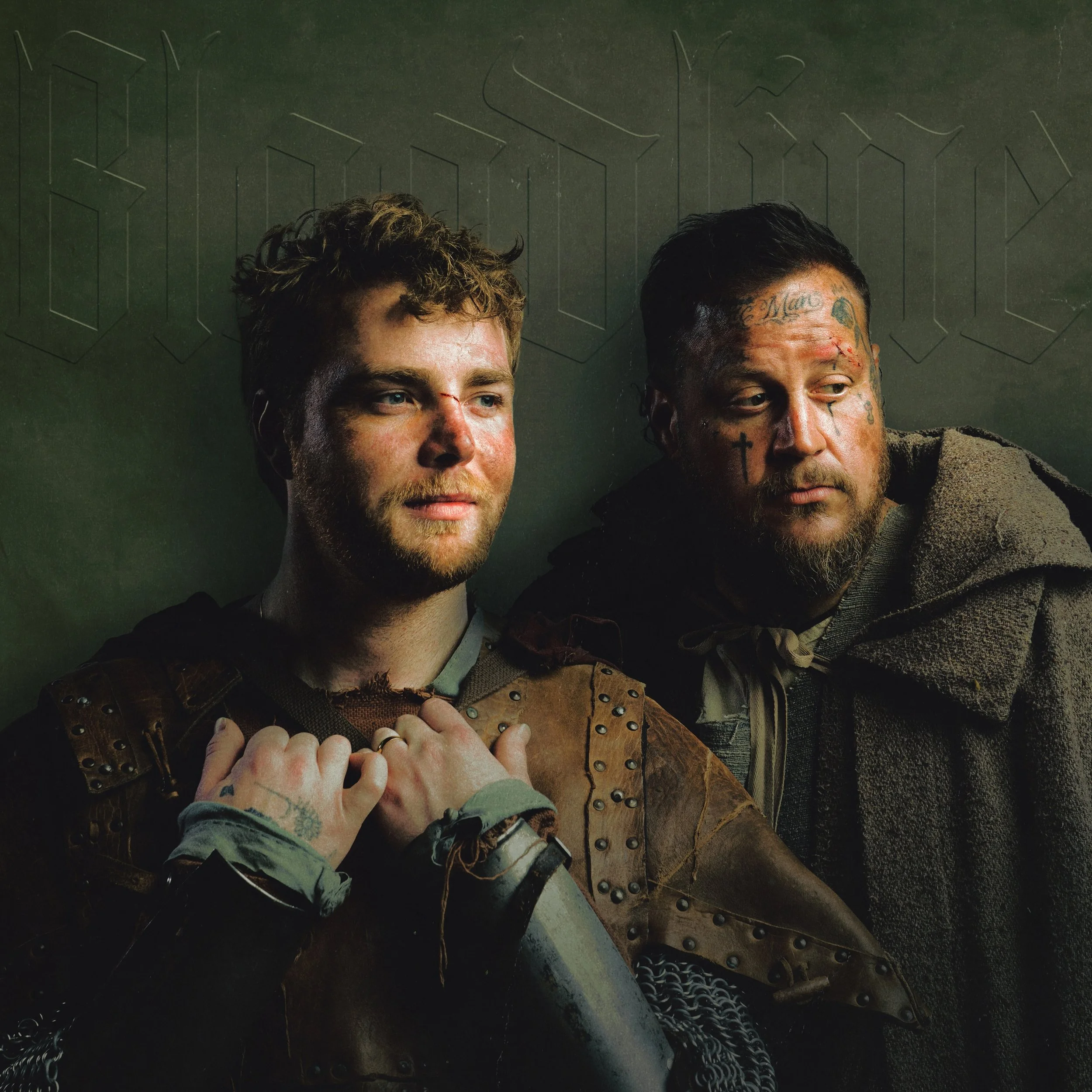 Two men with tattoos and piercings sitting close together, holding hands, against a dark background with a geometric pattern.