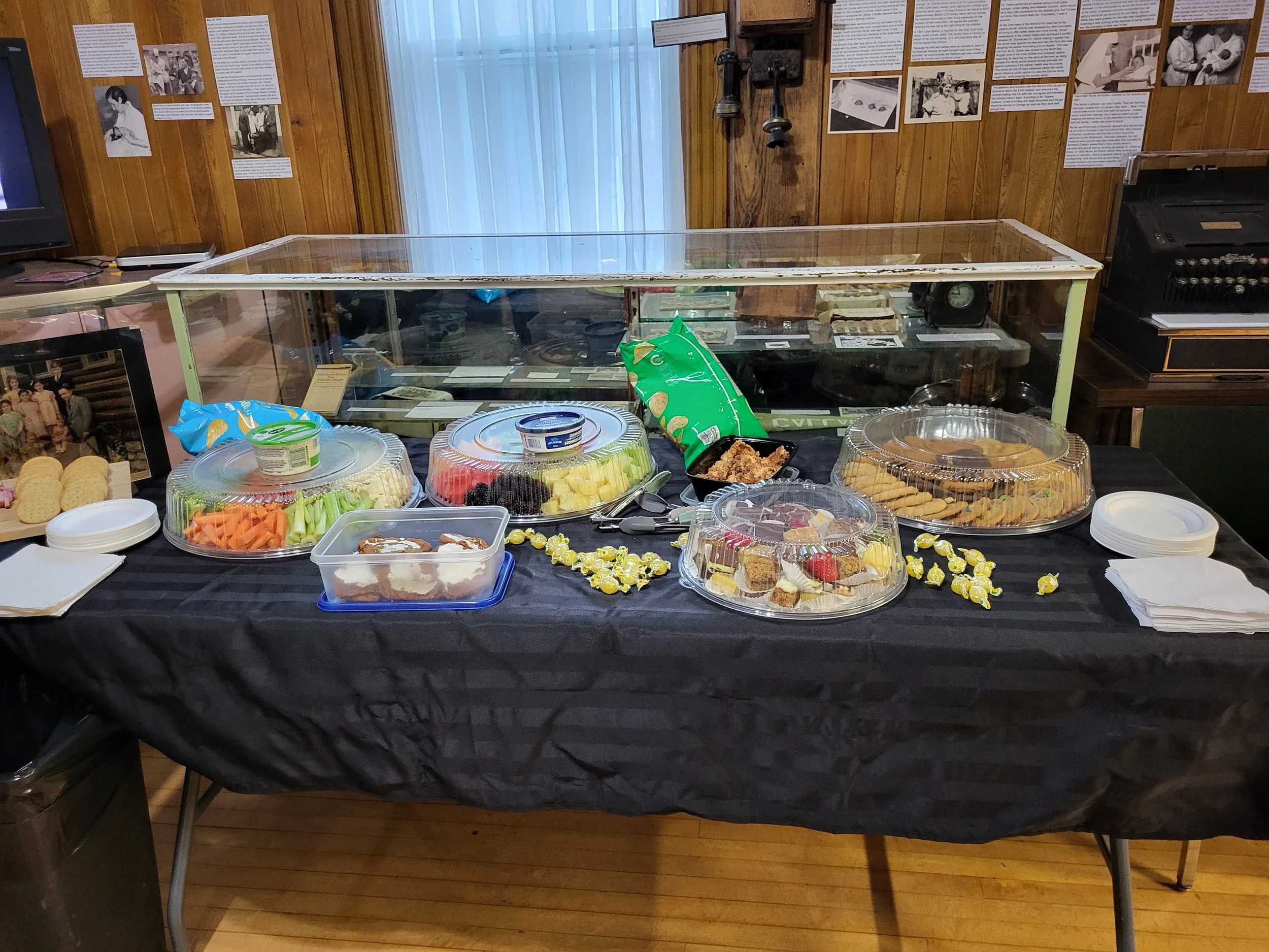 A display table of assorted snacks and desserts, including fruit, chips, cookies, and candies, in a room with wooden walls decorated with historical photographs and documents.