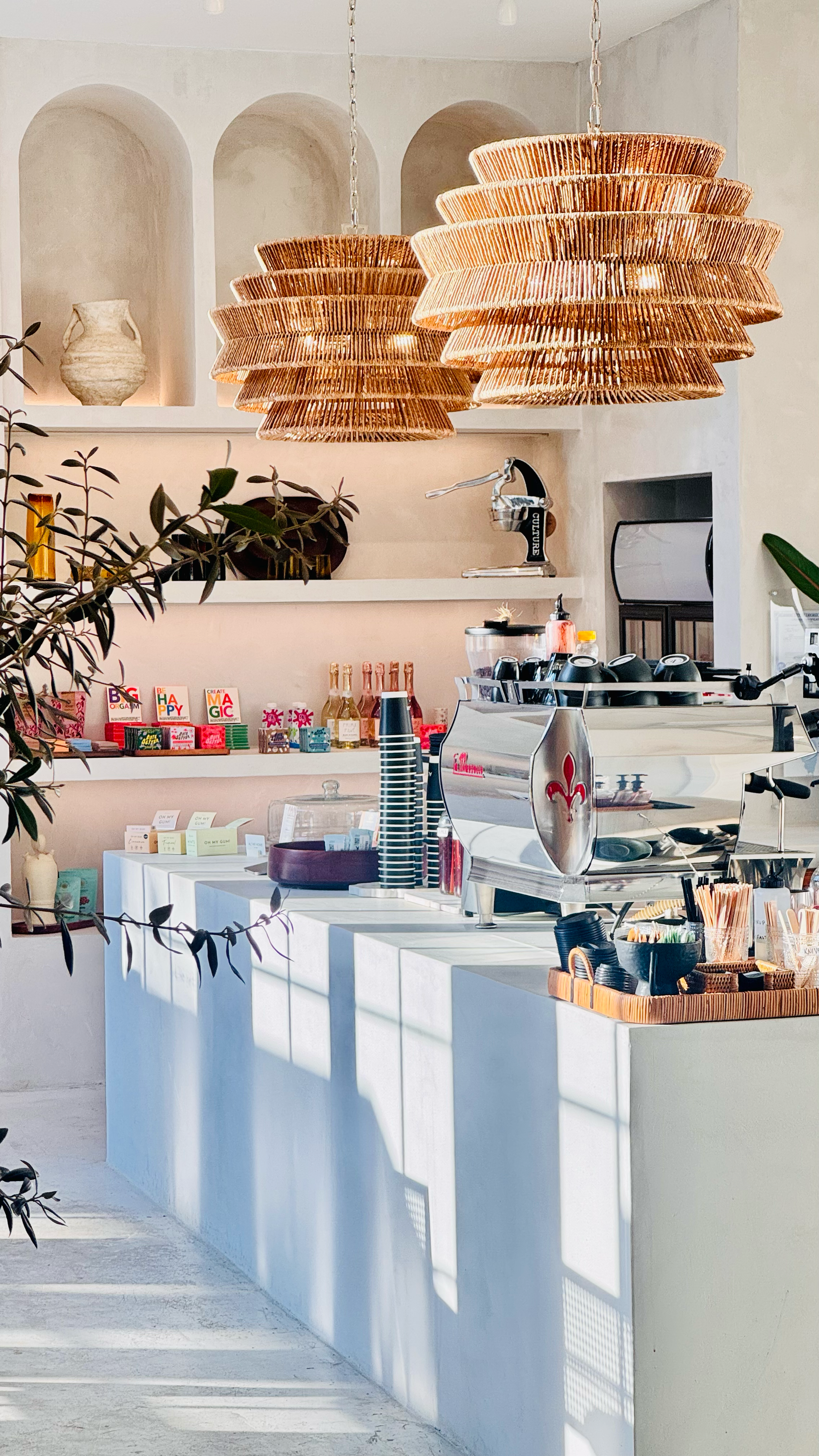 Interior view of a modern cafe or coffee shop with white countertops, hanging wicker pendant lights, a coffee machine, and shelves with colorful items and decorations.