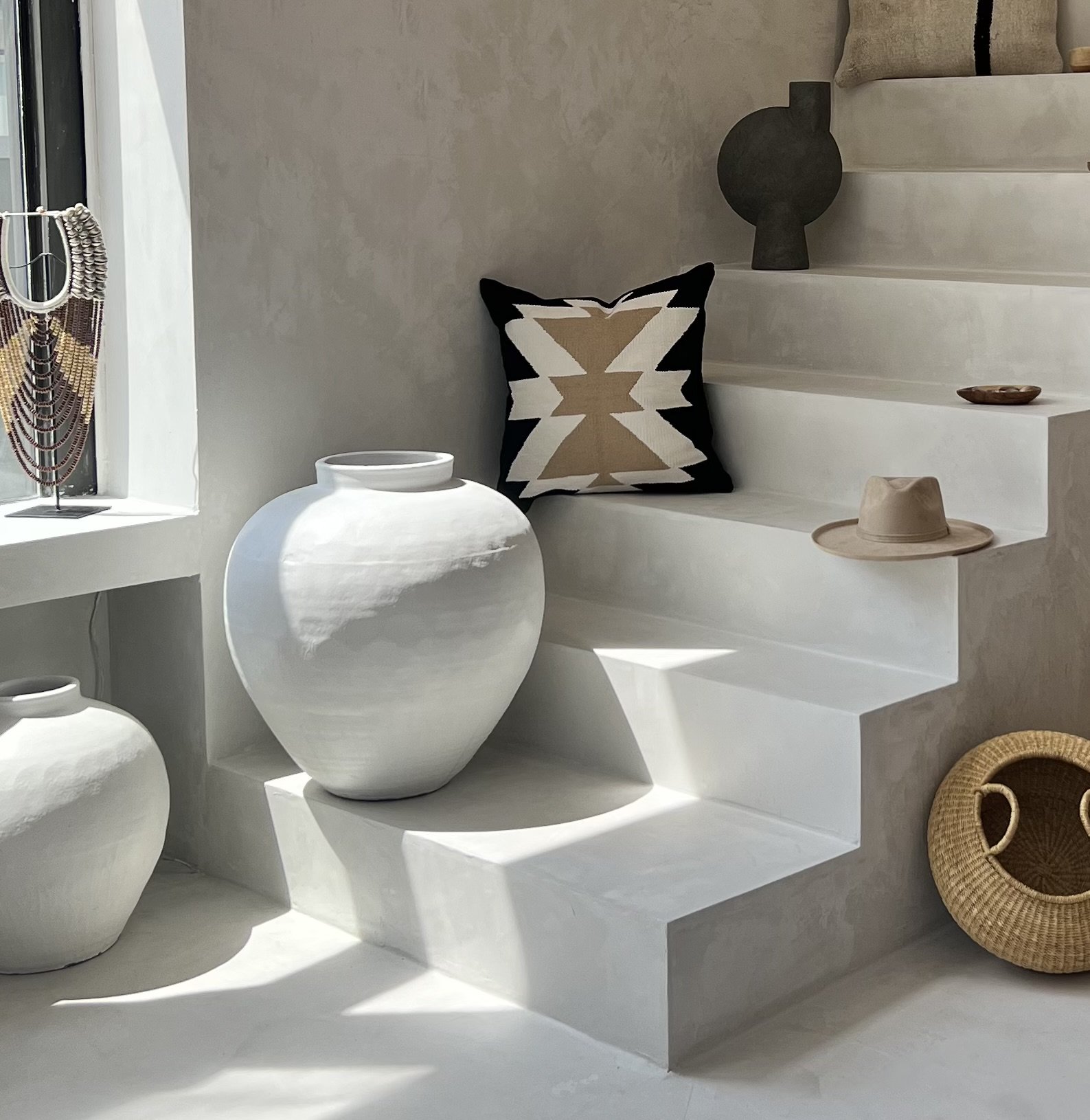 Minimalist interior with white ceramic vases, decorative pillows, woven basket, and neutral-toned accessories on stairs and window sill.