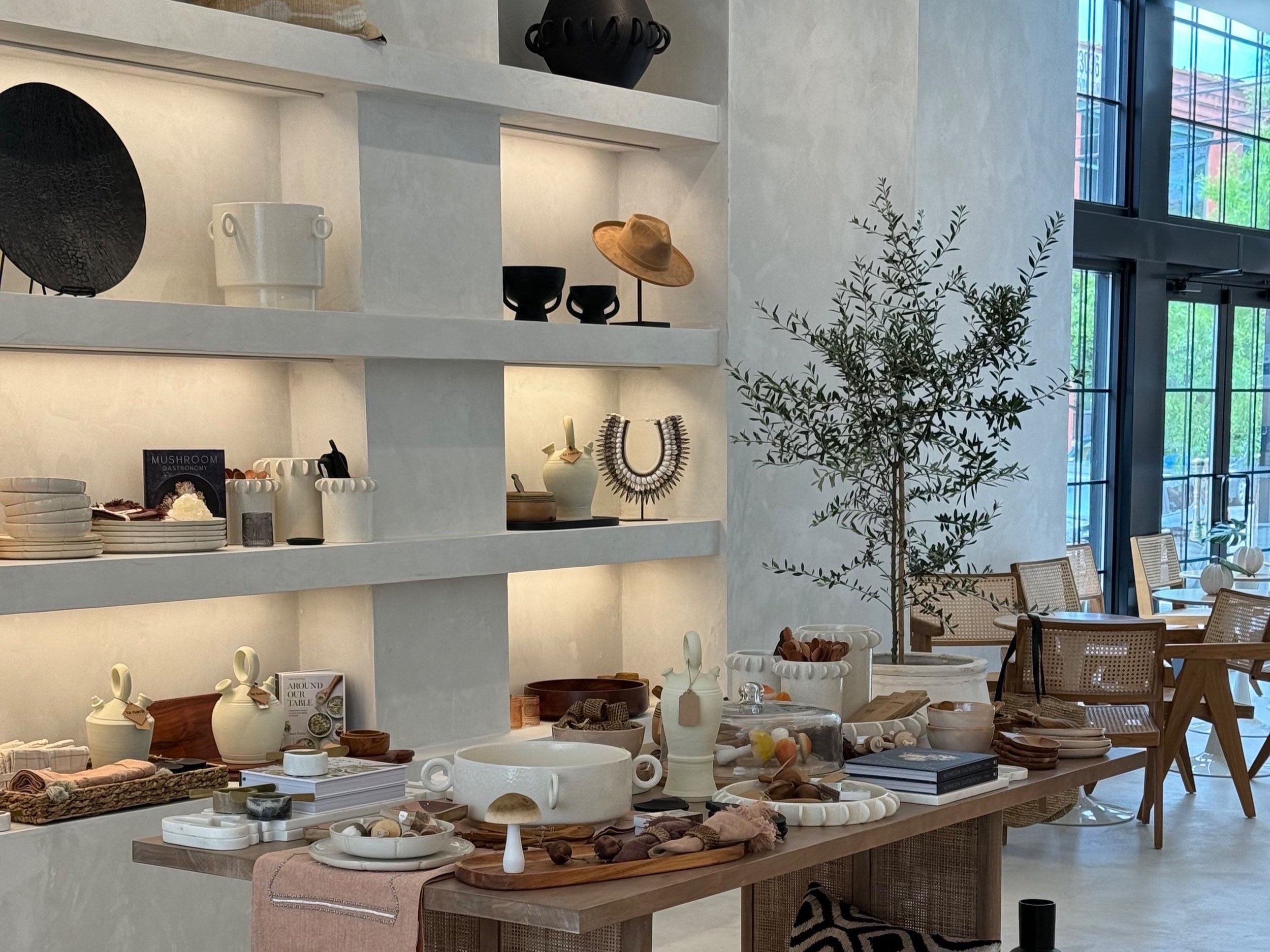 Interior of a modern store with various home decor and tableware items displayed on a wooden table and white shelves, next to large windows with outdoor greenery visible.