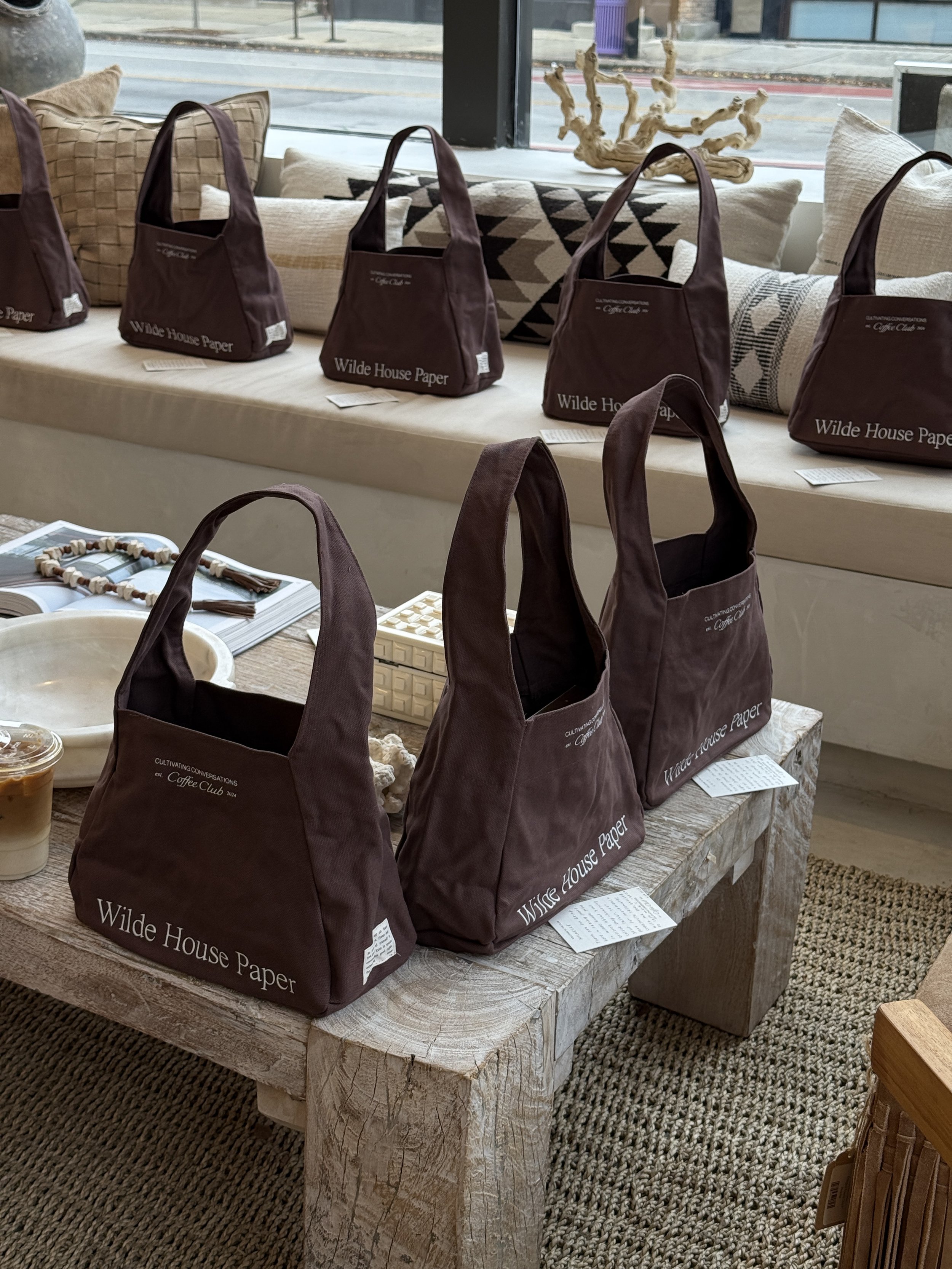 Multiple dark purple bags with white text on a wooden table in a retail store, with additional bags on a window seat and some decorative pillows in the background.
