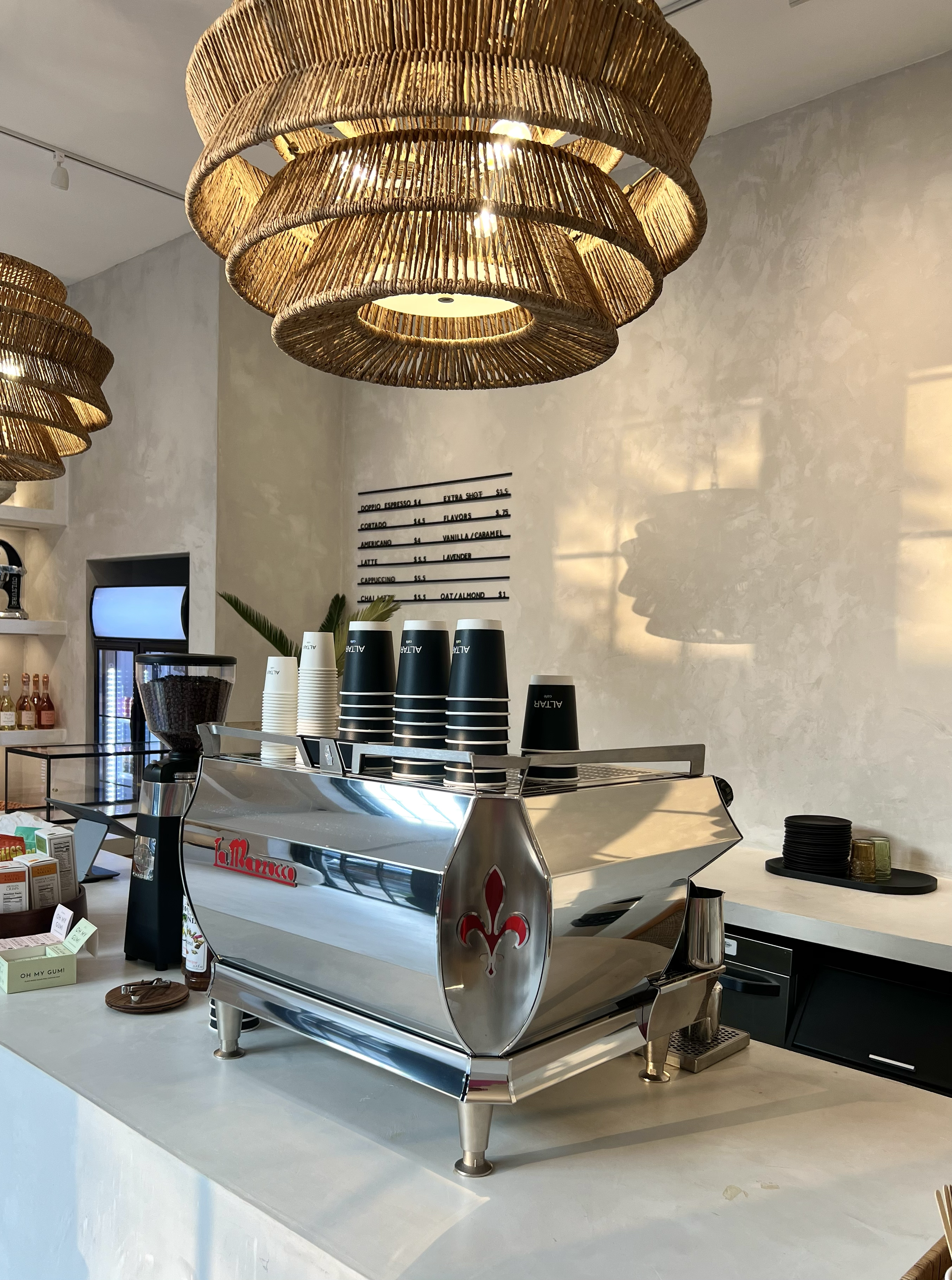 A modern coffee shop with a polished espresso machine on the counter, surrounded by cups and accessories, illuminated by large wooden pendant lights, with a menu board on the wall and decorative plants in the background.