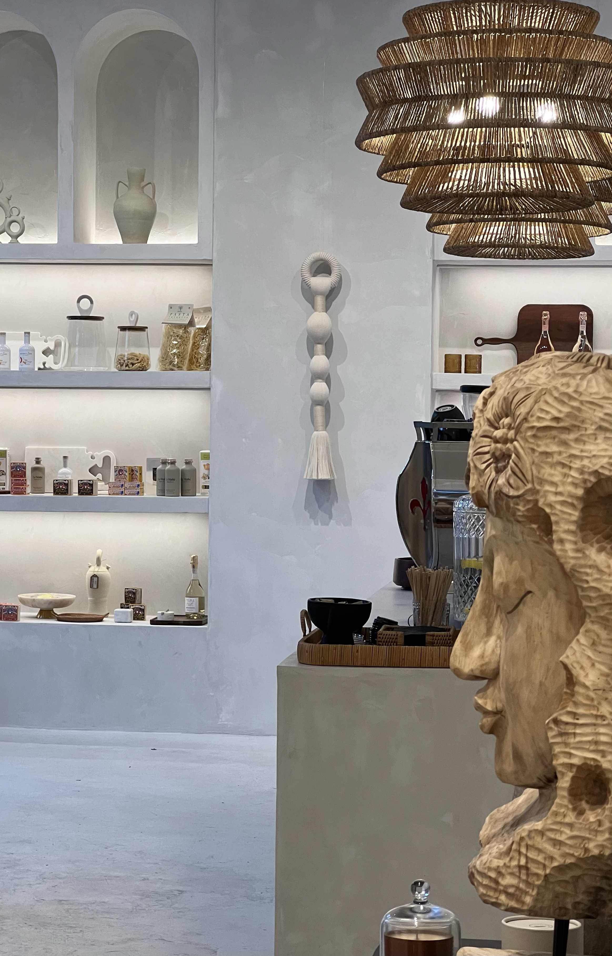 Interior of a minimalist boutique or cafe with decorative items on shelves, a hanging woven light fixture, and a wooden sculpture of a human face.