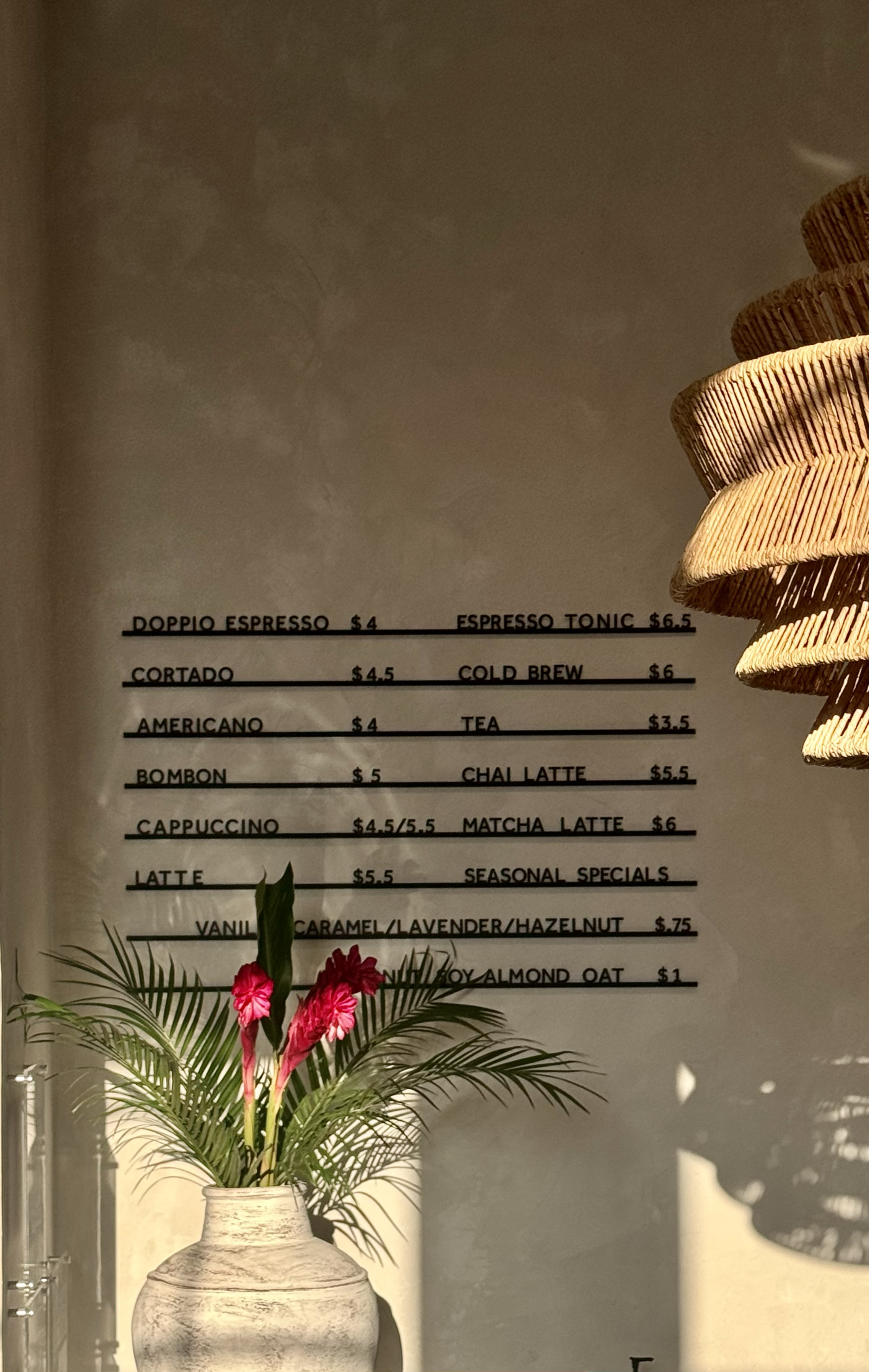 Cafe menu on the wall with coffee and tea options, priced between $3.5 and $6.5, with a vase holding pink flowers and green foliage in the foreground.