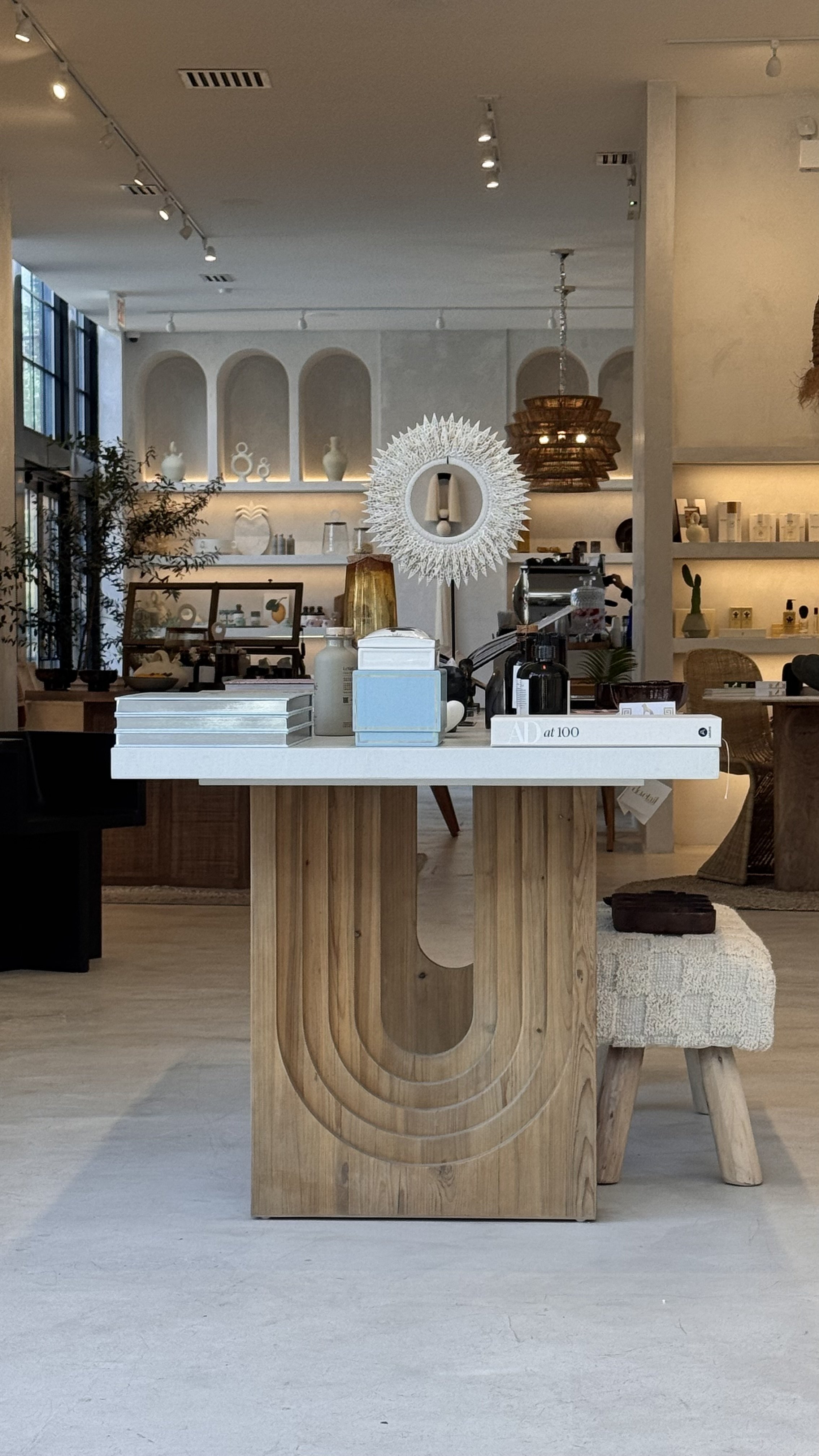 A retail store display table made of wood with a white top, decorated with vases, books, and small items, in a modern interior with shelves and decorative objects in the background.